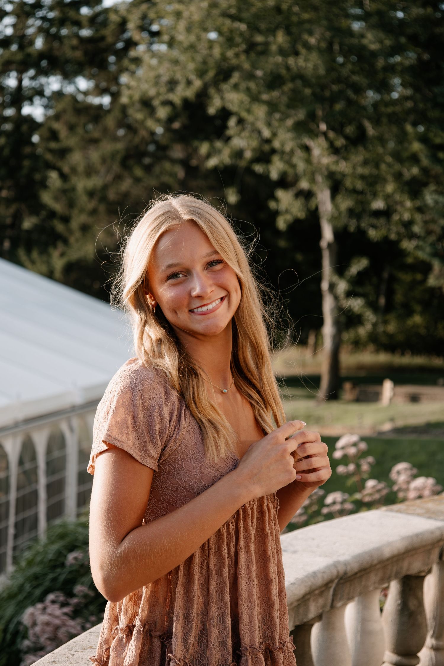 Smiling blonde woman in a pink floral dress standing on a stone balcony with lush green trees in the background.