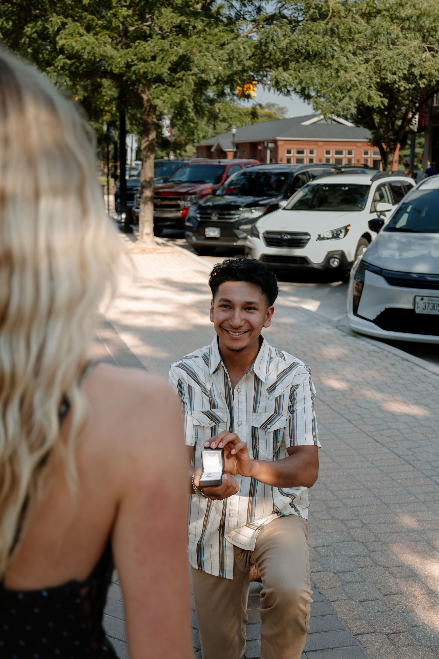 Man on one knee proposing with a ring box to a blonde woman on a brick sidewalk outdoors.