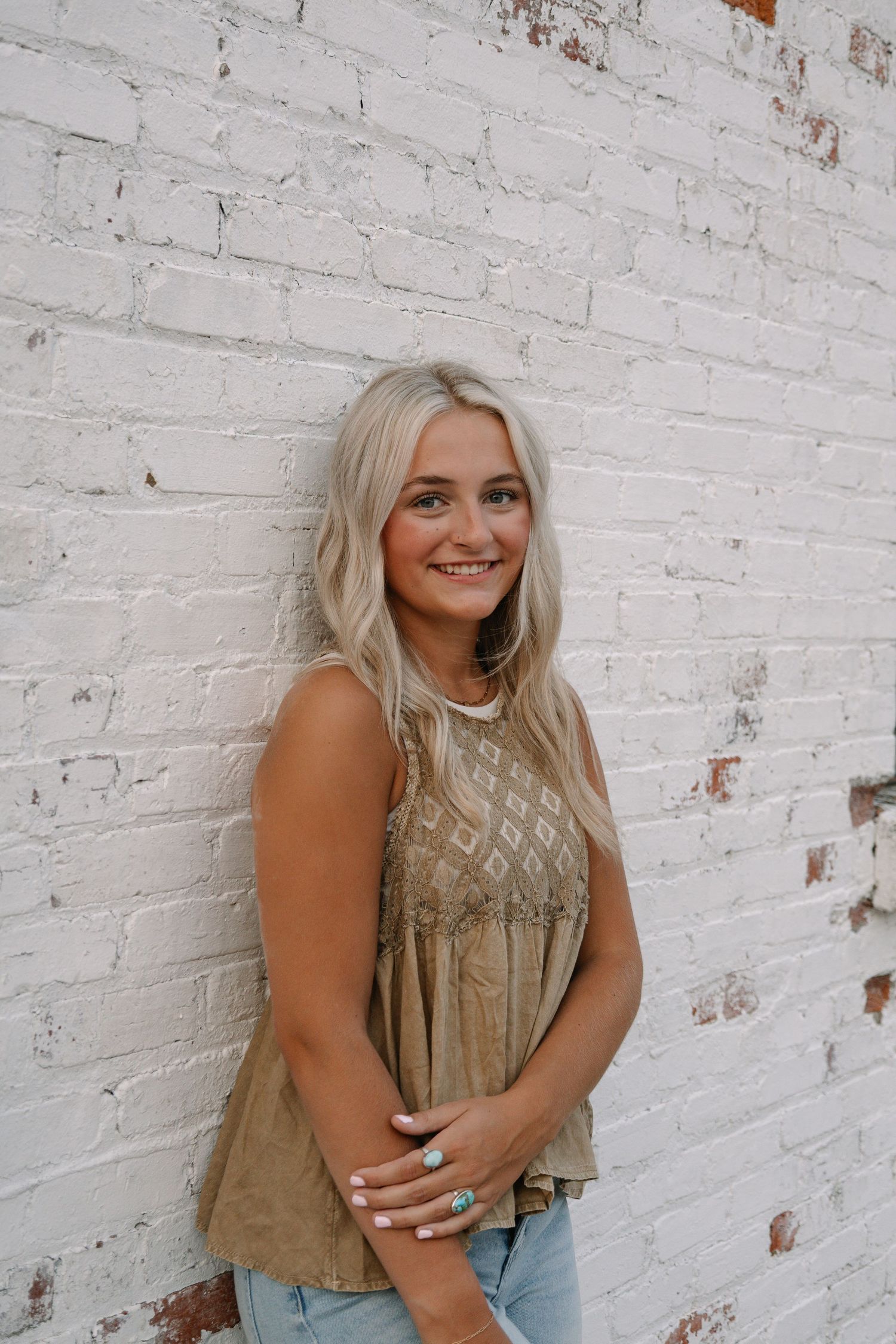Smiling blonde woman in olive sleeveless top leaning against white brick wall wearing turquoise ring.