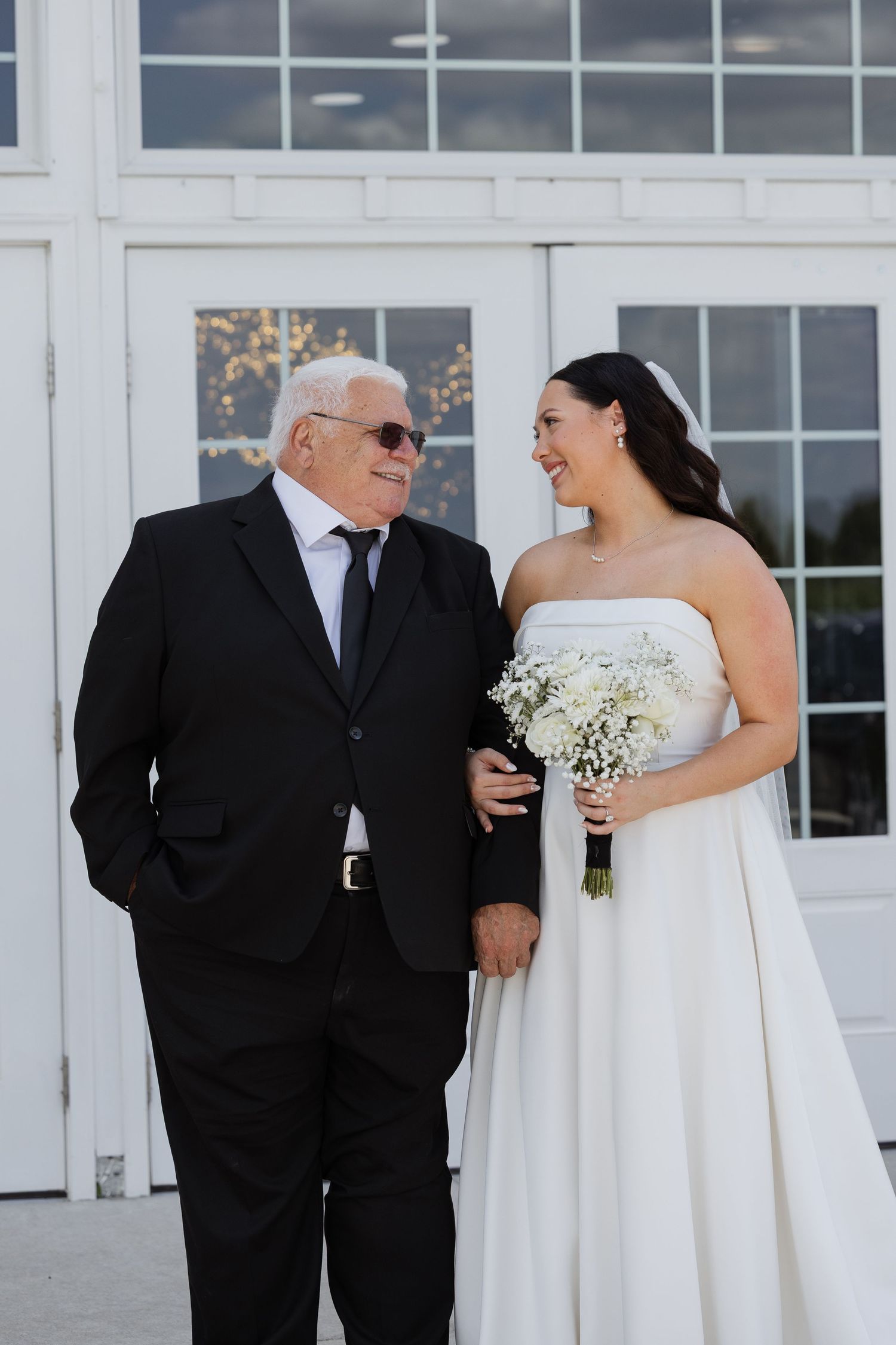 Bride in strapless white gown holding bouquet smiles at elderly man in black suit outside white venue.