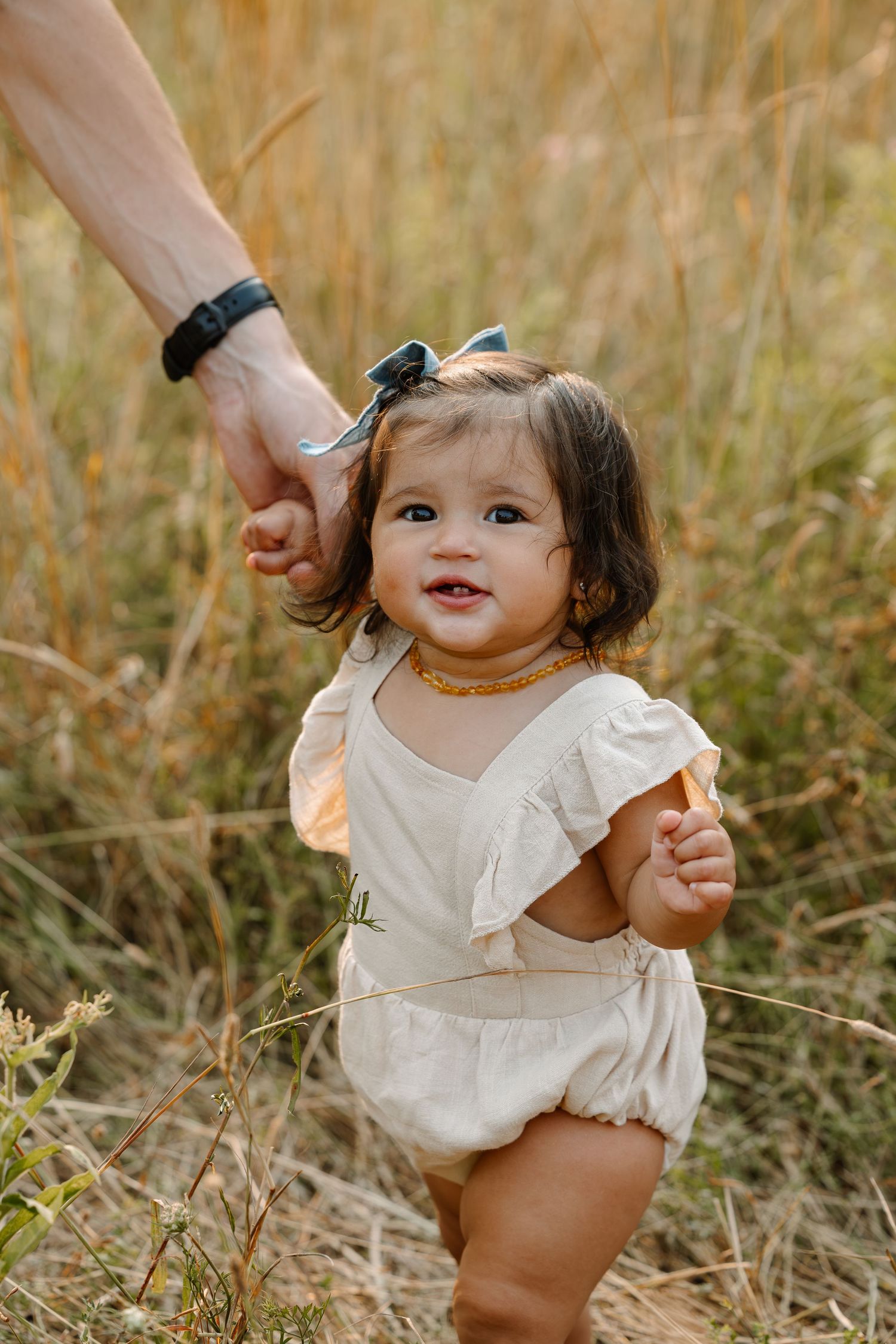 Smiling toddler girl in cream romper holding adult hand while standing in golden field of tall grass.