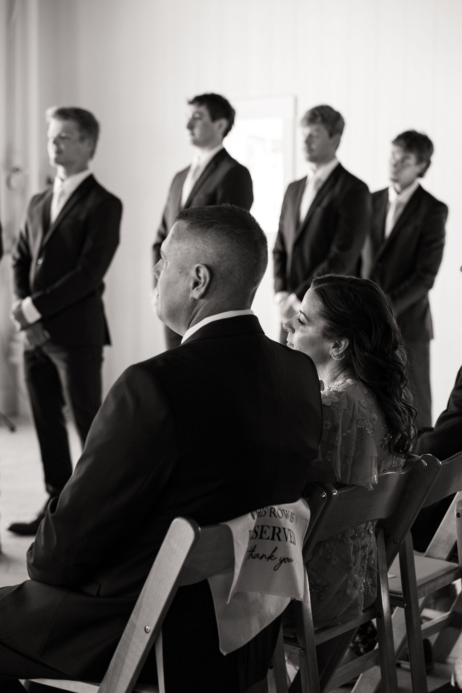Black and white photo of wedding guests seated while groomsmen stand at altar ceremony.