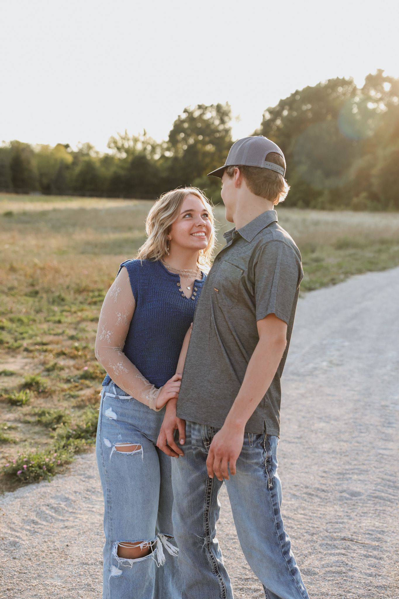 A couple walks along a sunny gravel path, smiling at each other, surrounded by golden fields at sunset.