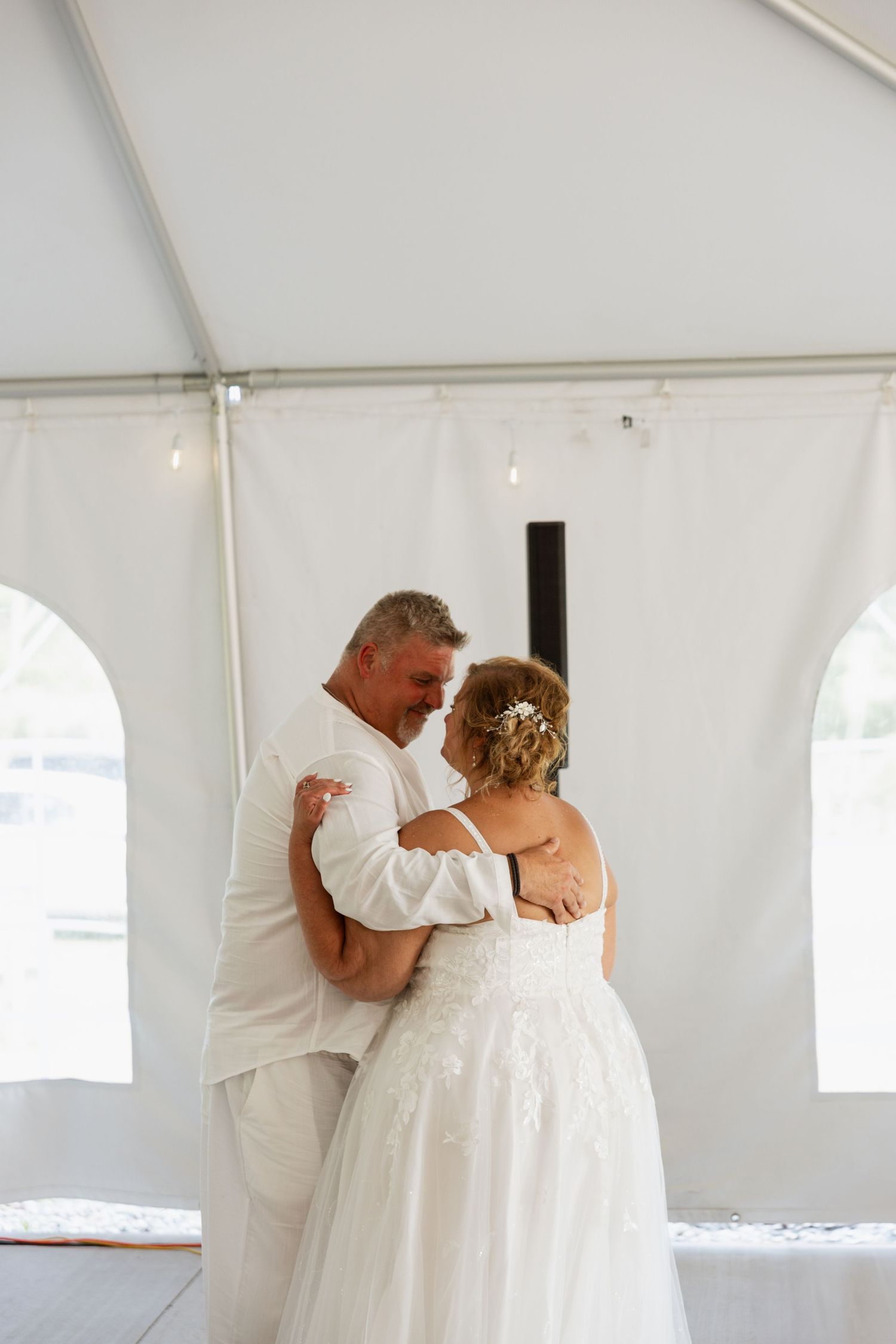 Newlywed couple shares intimate first dance embrace under white tent at outdoor wedding reception.