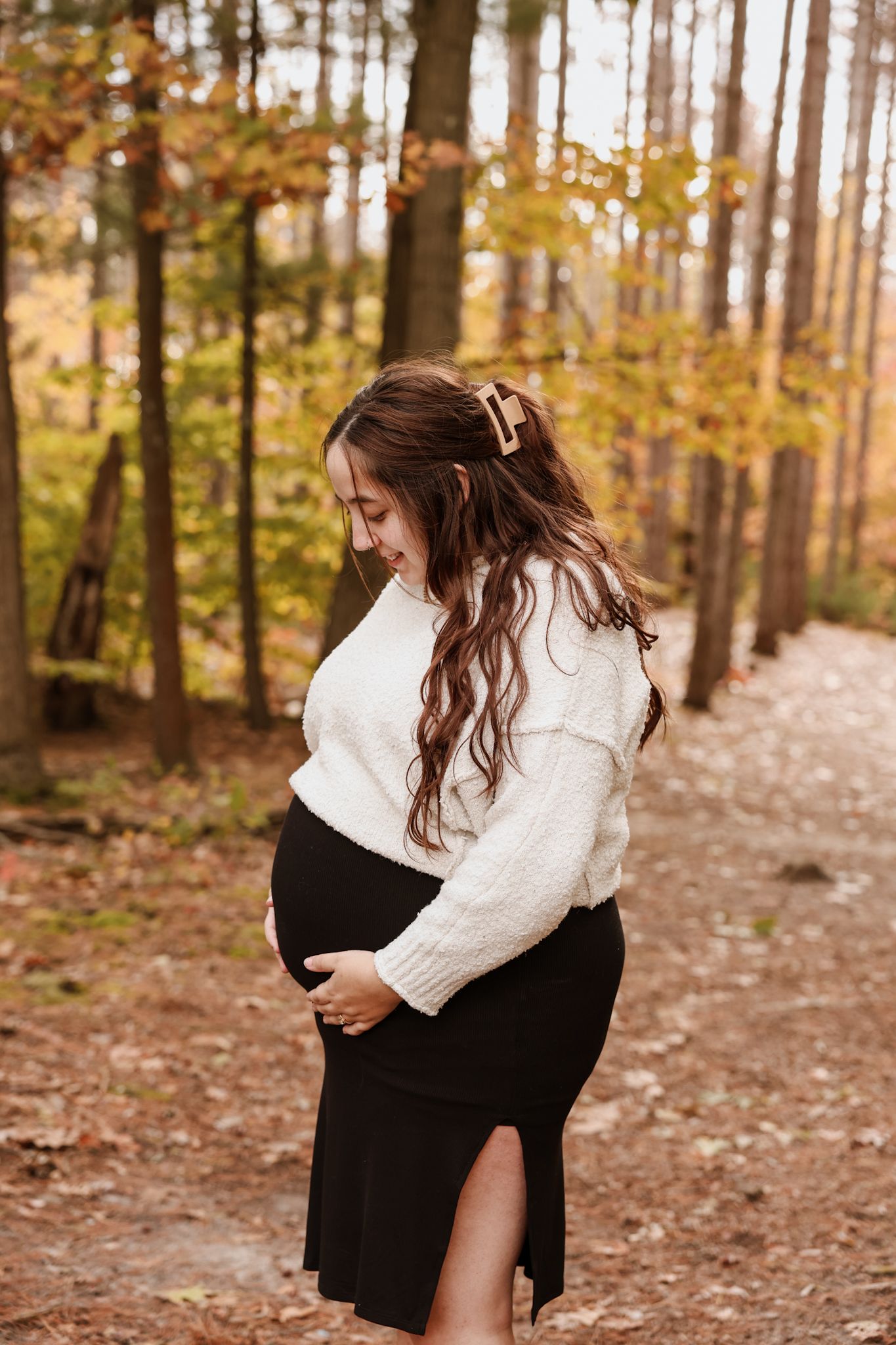 Pregnant woman in white sweater and black skirt cradling her baby bump in an autumn forest.