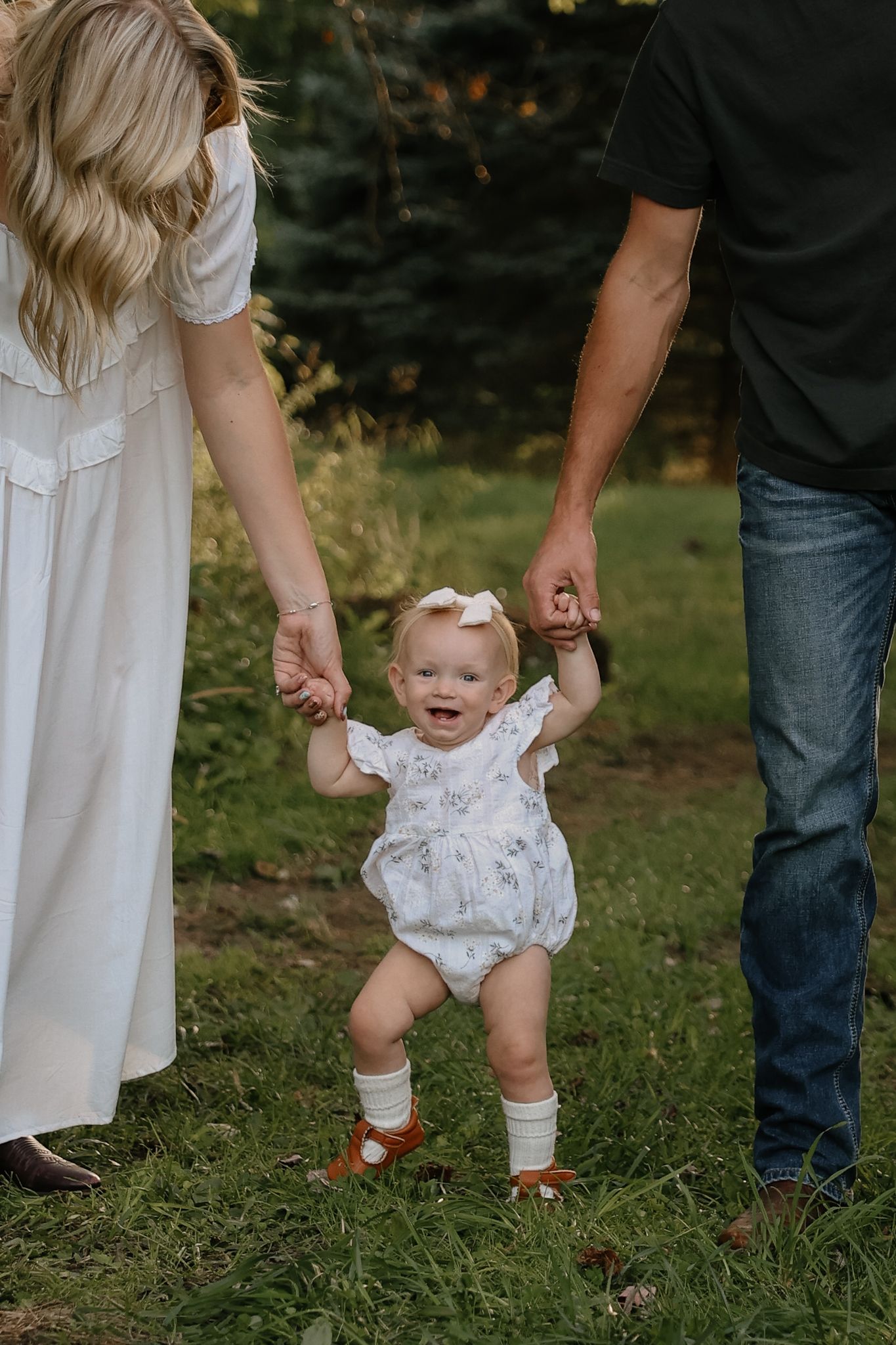 Baby girl in floral romper taking first steps while holding parents hands in green outdoor setting.
