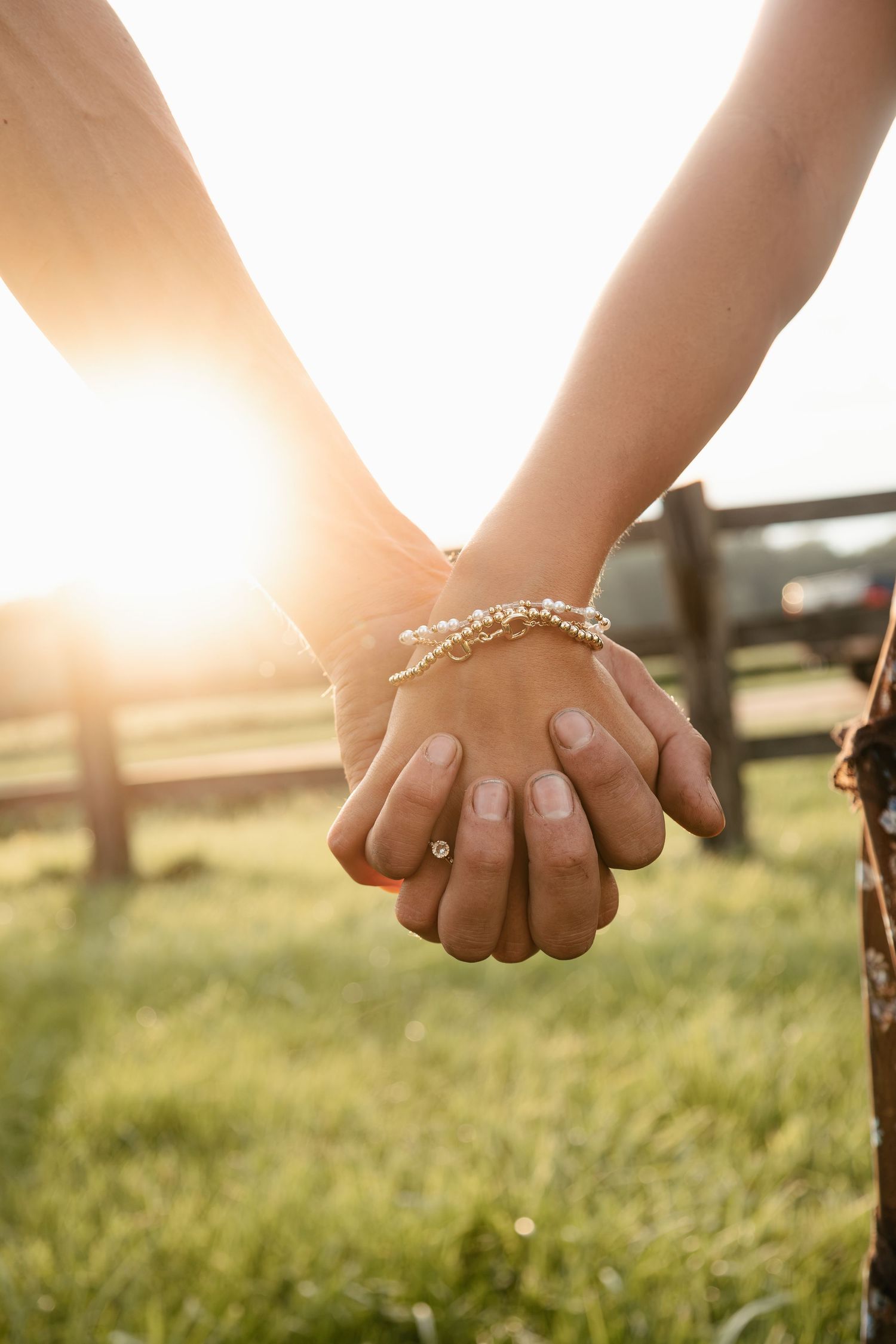Two people holding hands at sunset in a green field, one wearing a delicate bracelet and a ring.