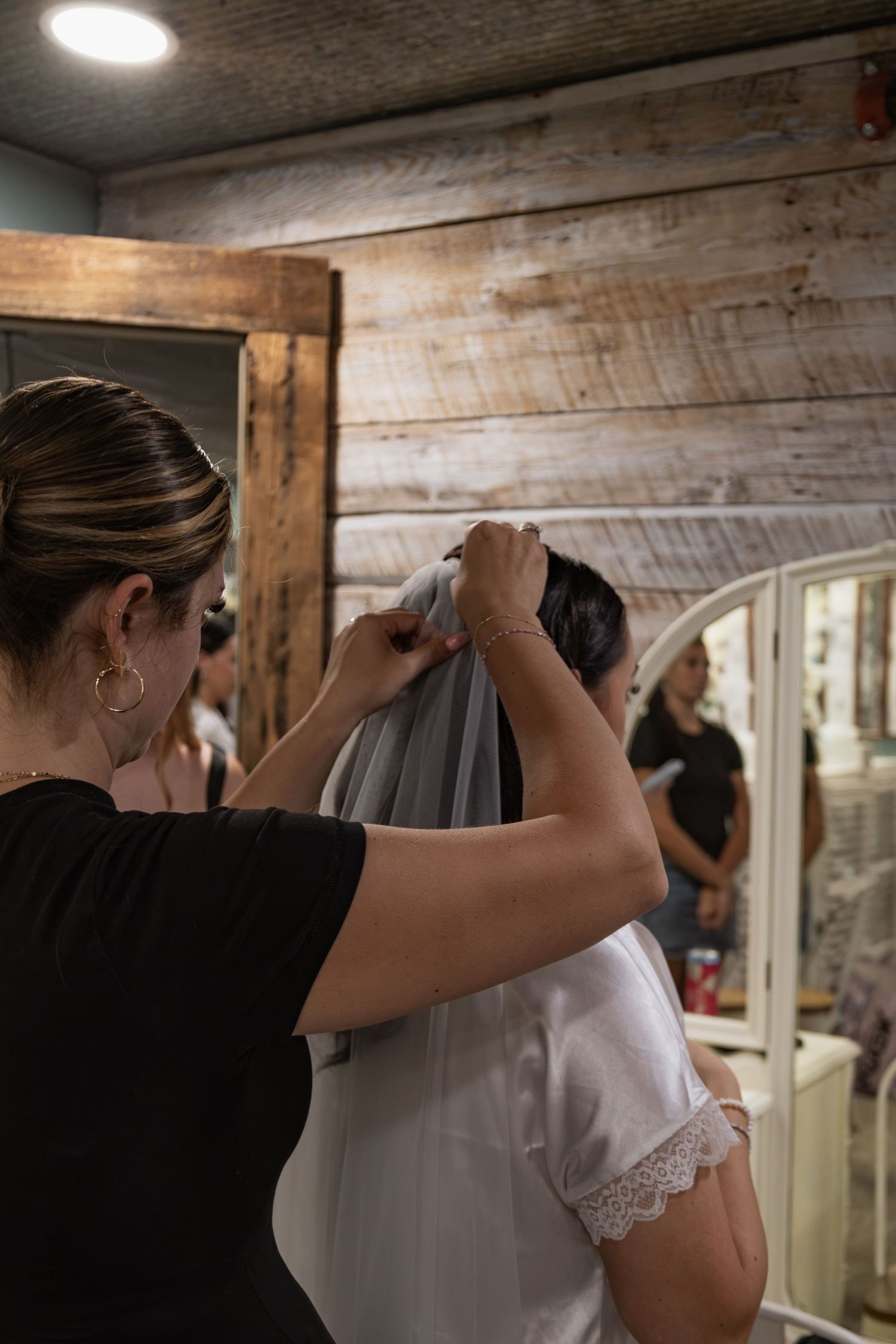 Bridesmaid helping bride attach veil to hair in rustic wooden dressing room before wedding ceremony.