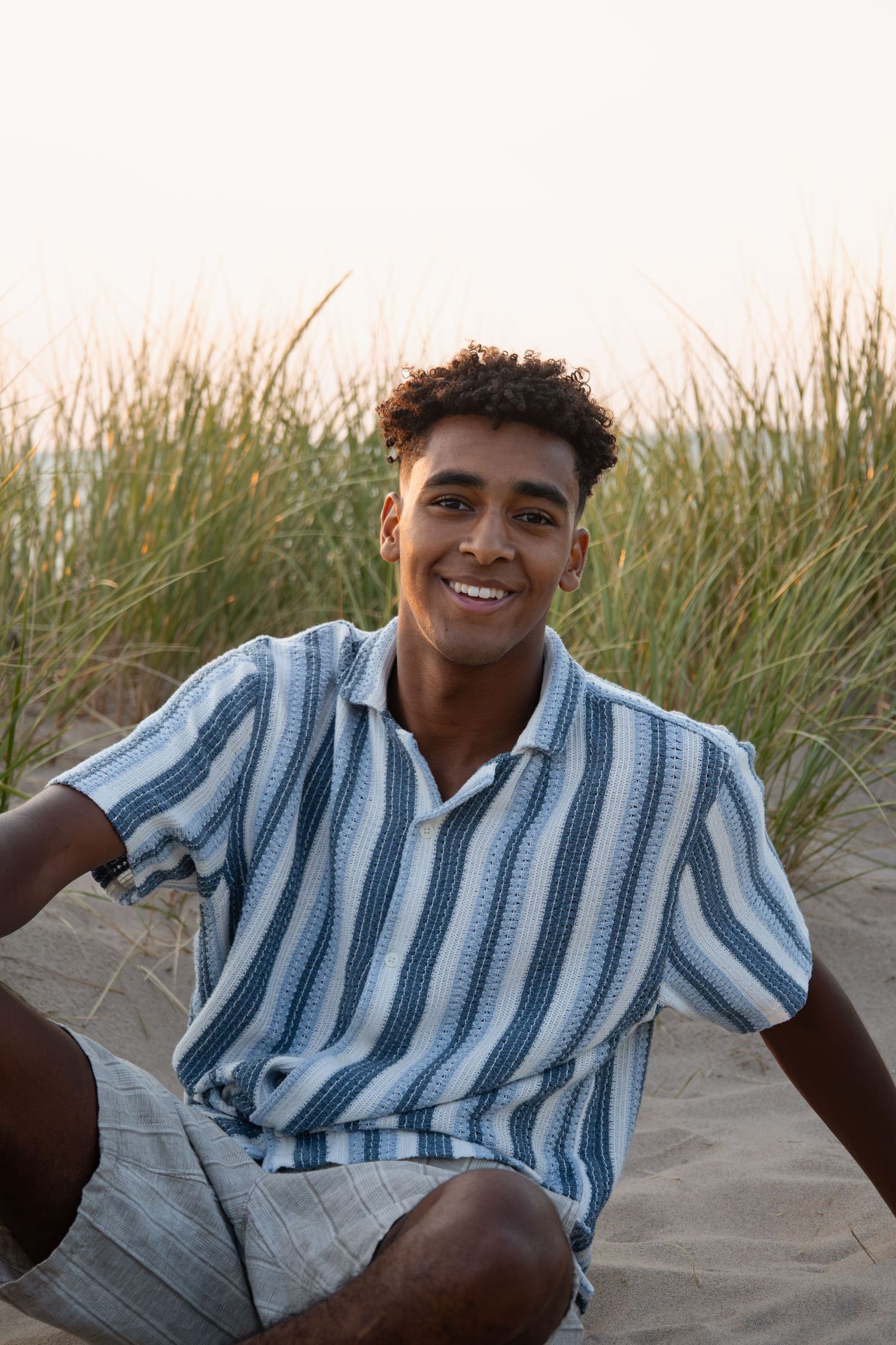 Young man in striped shirt sitting on sandy beach dunes with green grass, smiling at camera during golden hour.