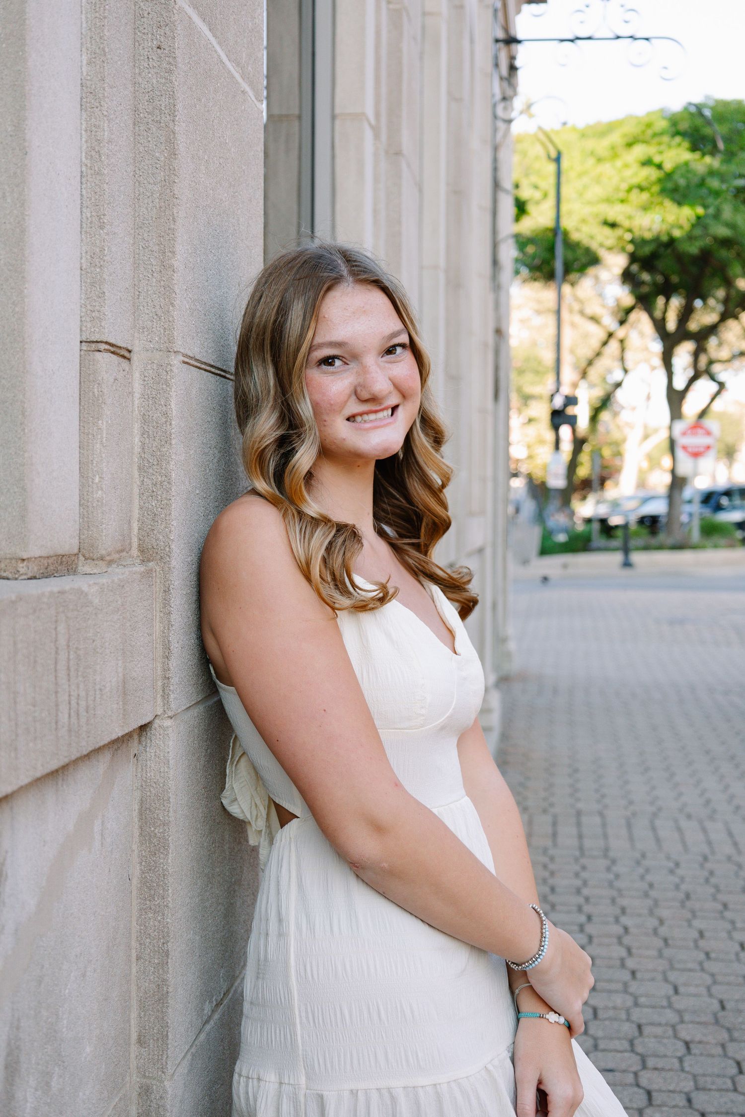 Young woman in white dress leaning against stone wall, smiling outdoors on urban sidewalk with trees in background.