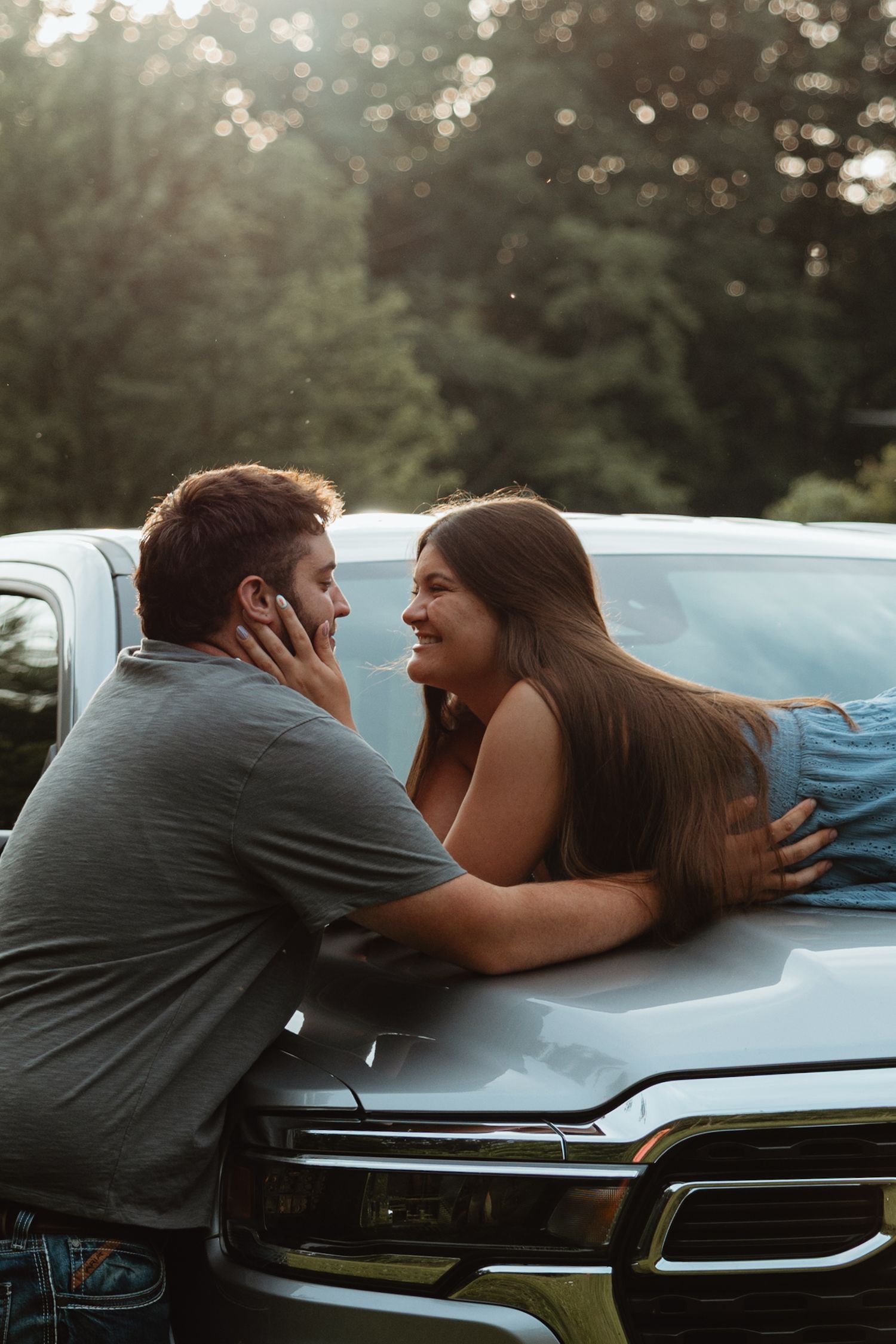 Couple sharing a romantic moment leaning against a silver truck hood in a sunlit forest setting.
