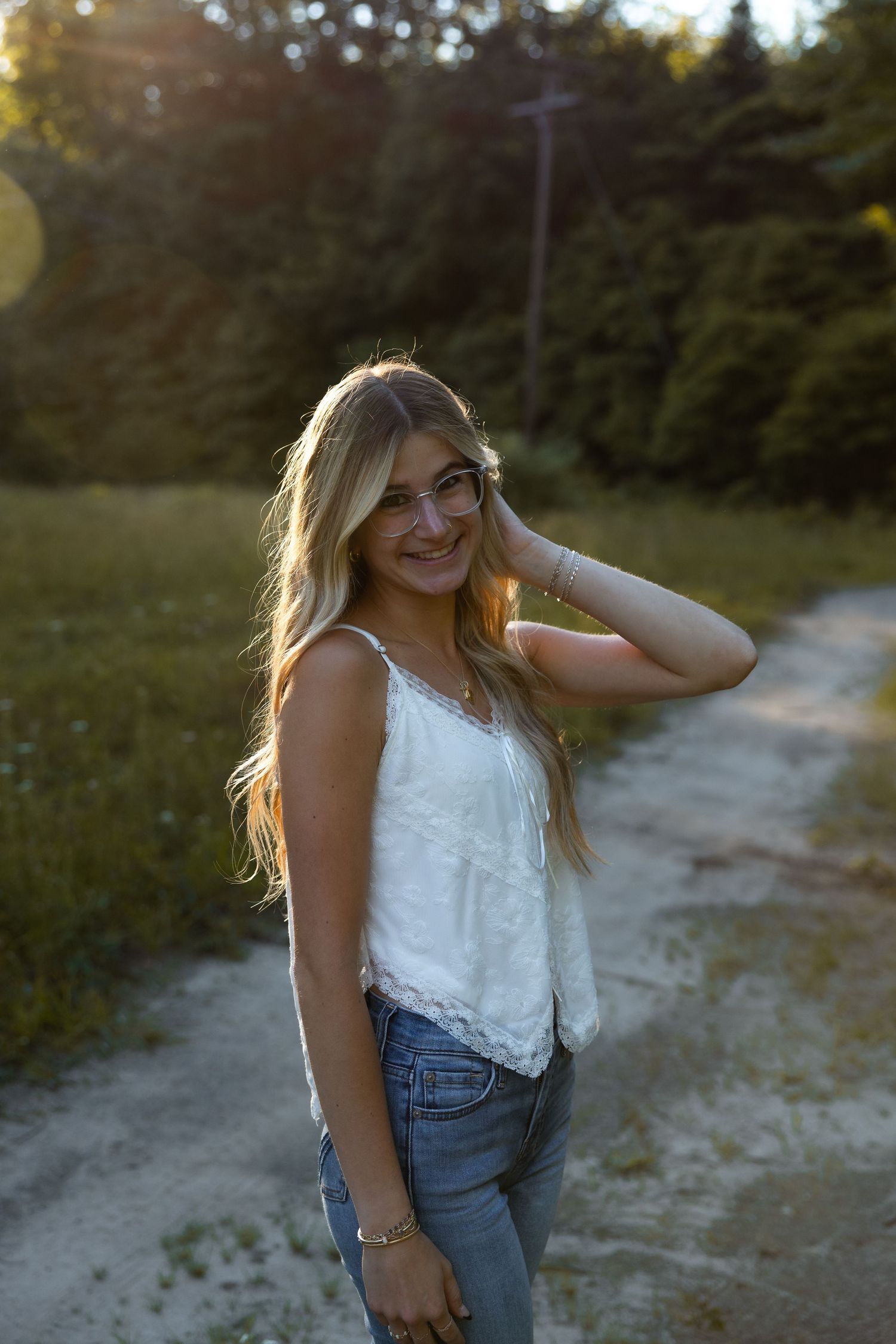Smiling young blonde woman in white lace tank top and jeans posing on a dirt path at golden hour sunset.