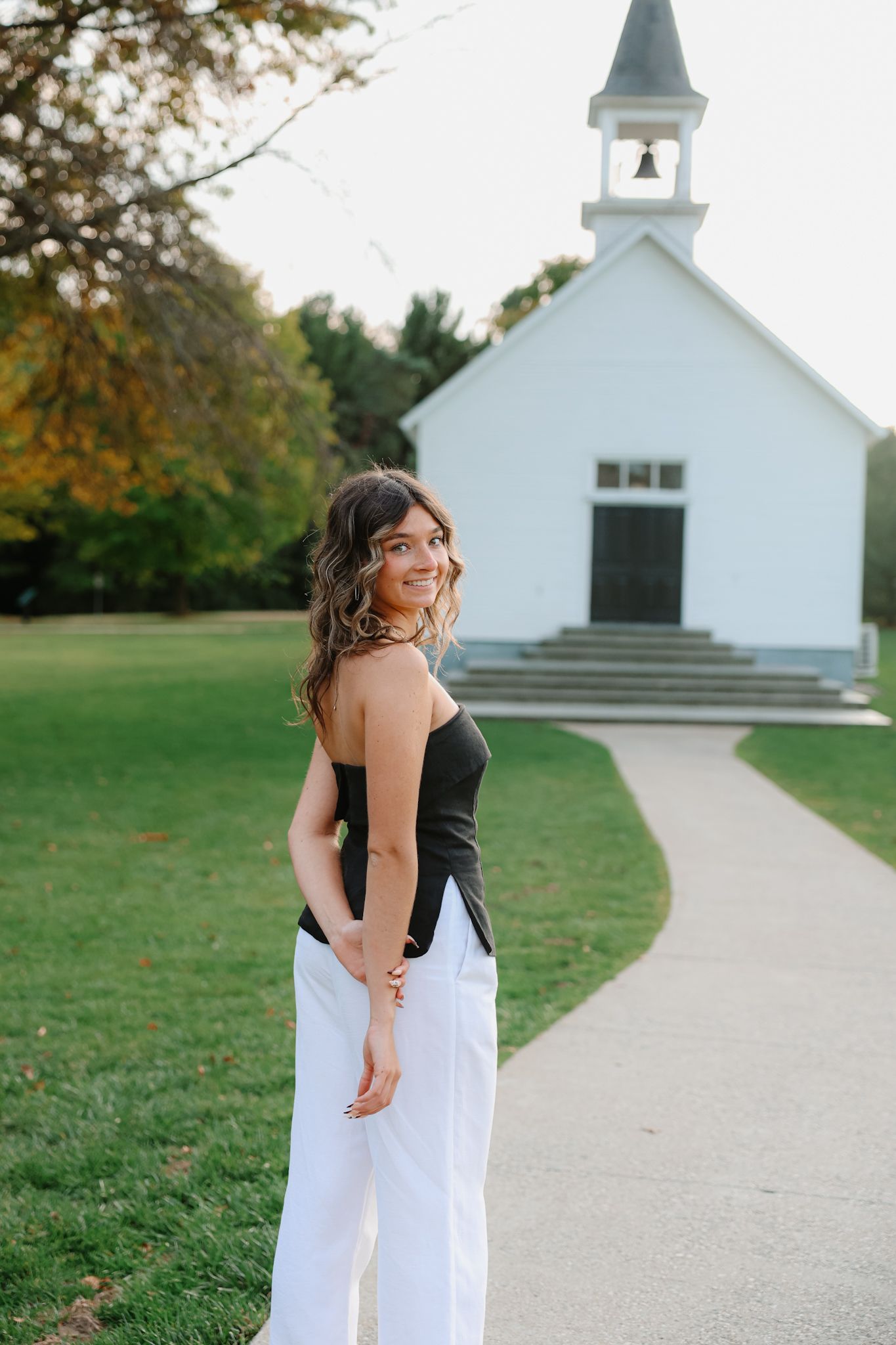 Young woman in black top and white pants smiling over shoulder near a white chapel on a sunny day.