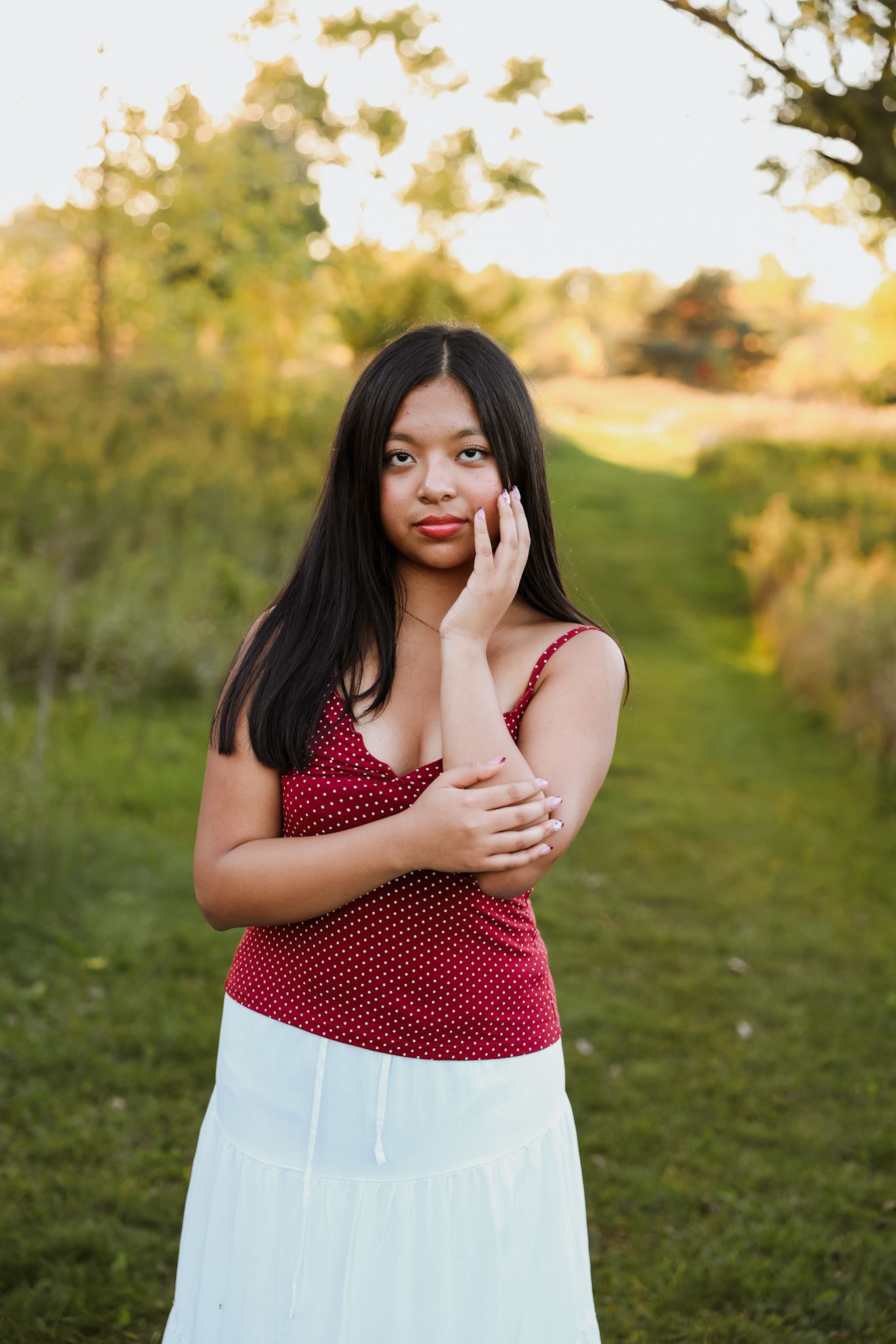 Young Asian woman in red polka dot top and white skirt poses thoughtfully in a green park setting.