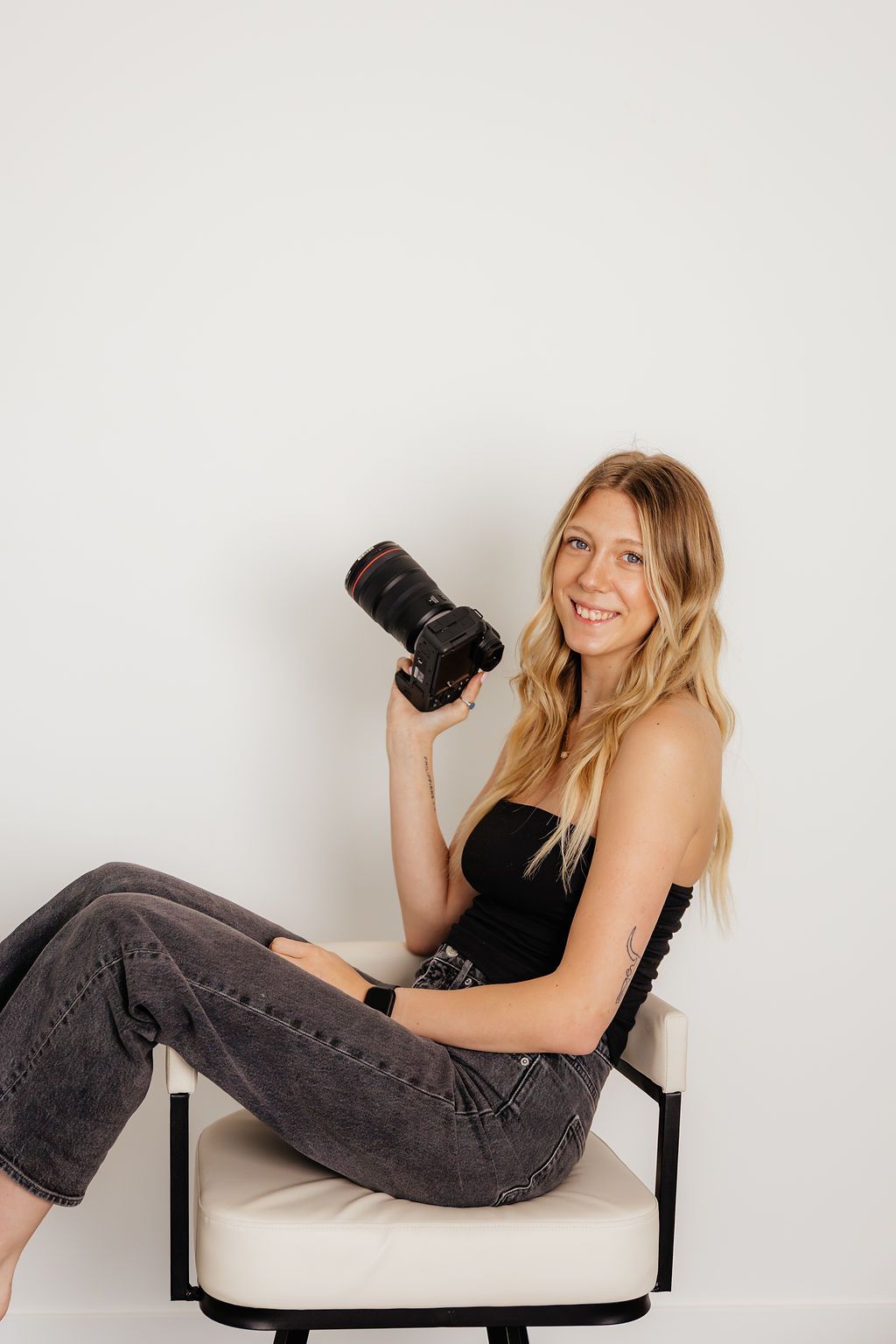 Smiling blonde female photographer seated in white chair holding DSLR camera wearing black tube top and gray jeans.