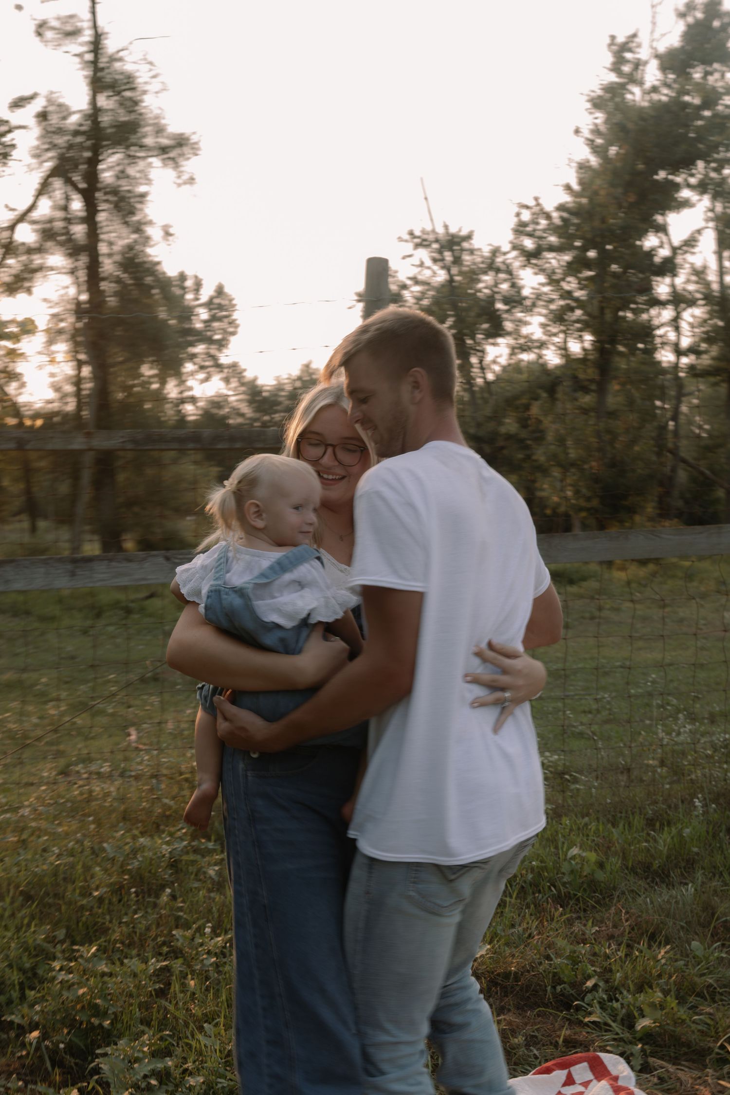 A young family shares a tender moment outdoors at sunset, with parents embracing while holding their blonde toddler.