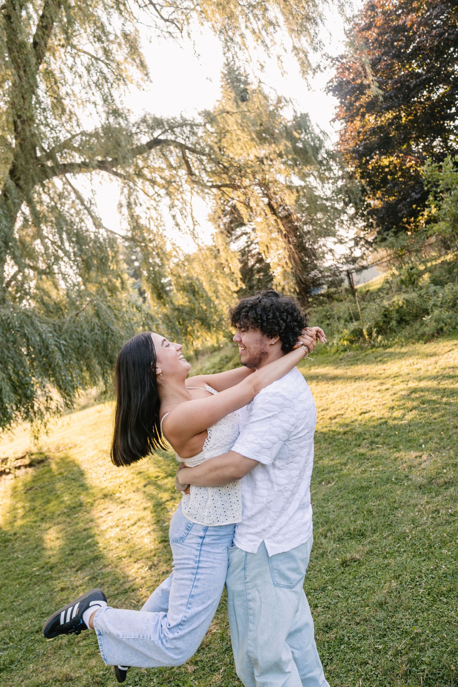 A joyful couple embraces and dances together in a sunny park under a weeping willow tree.