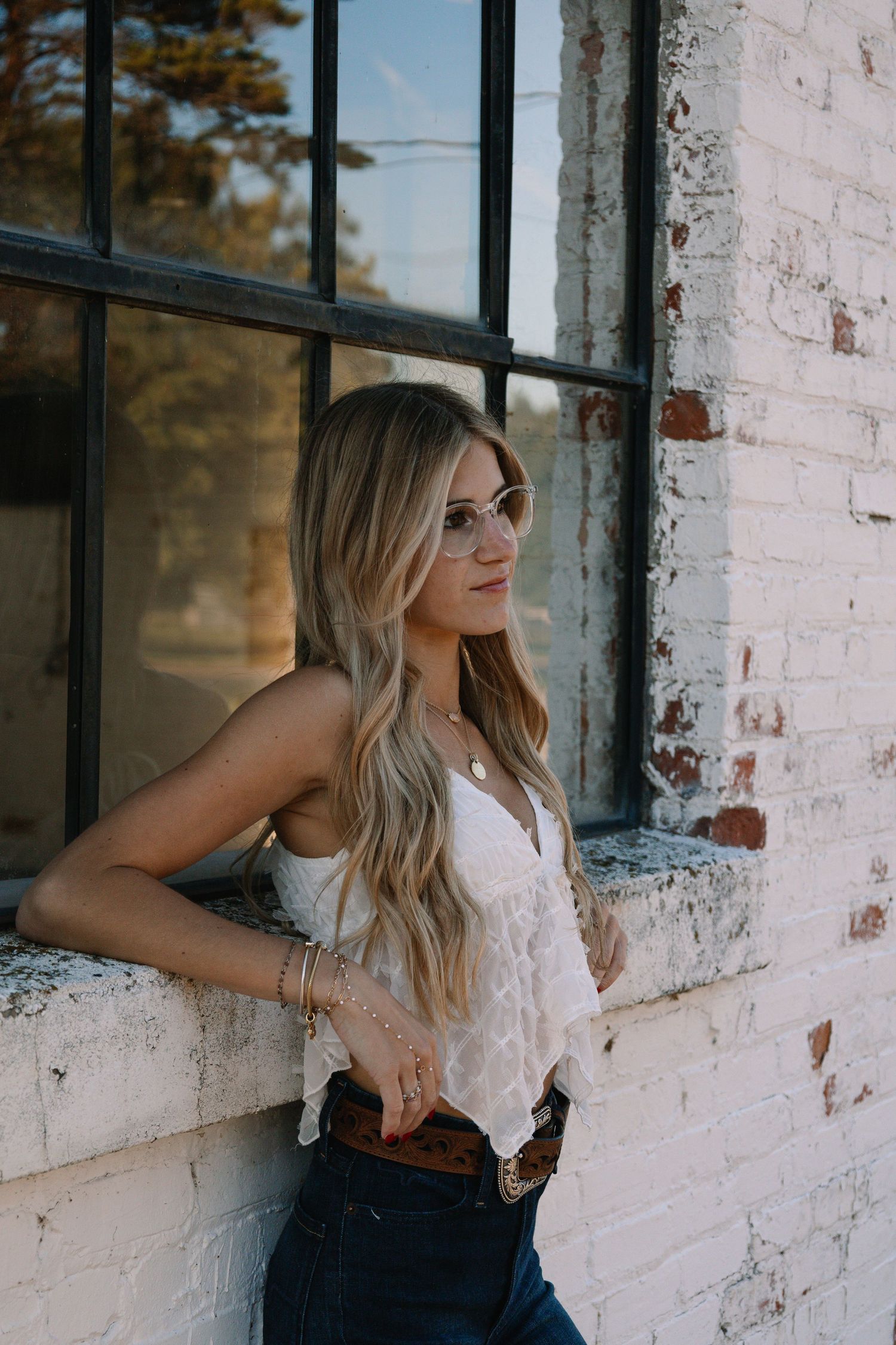 Blonde woman in white lace top and jeans leaning against a rustic brick wall by a window.