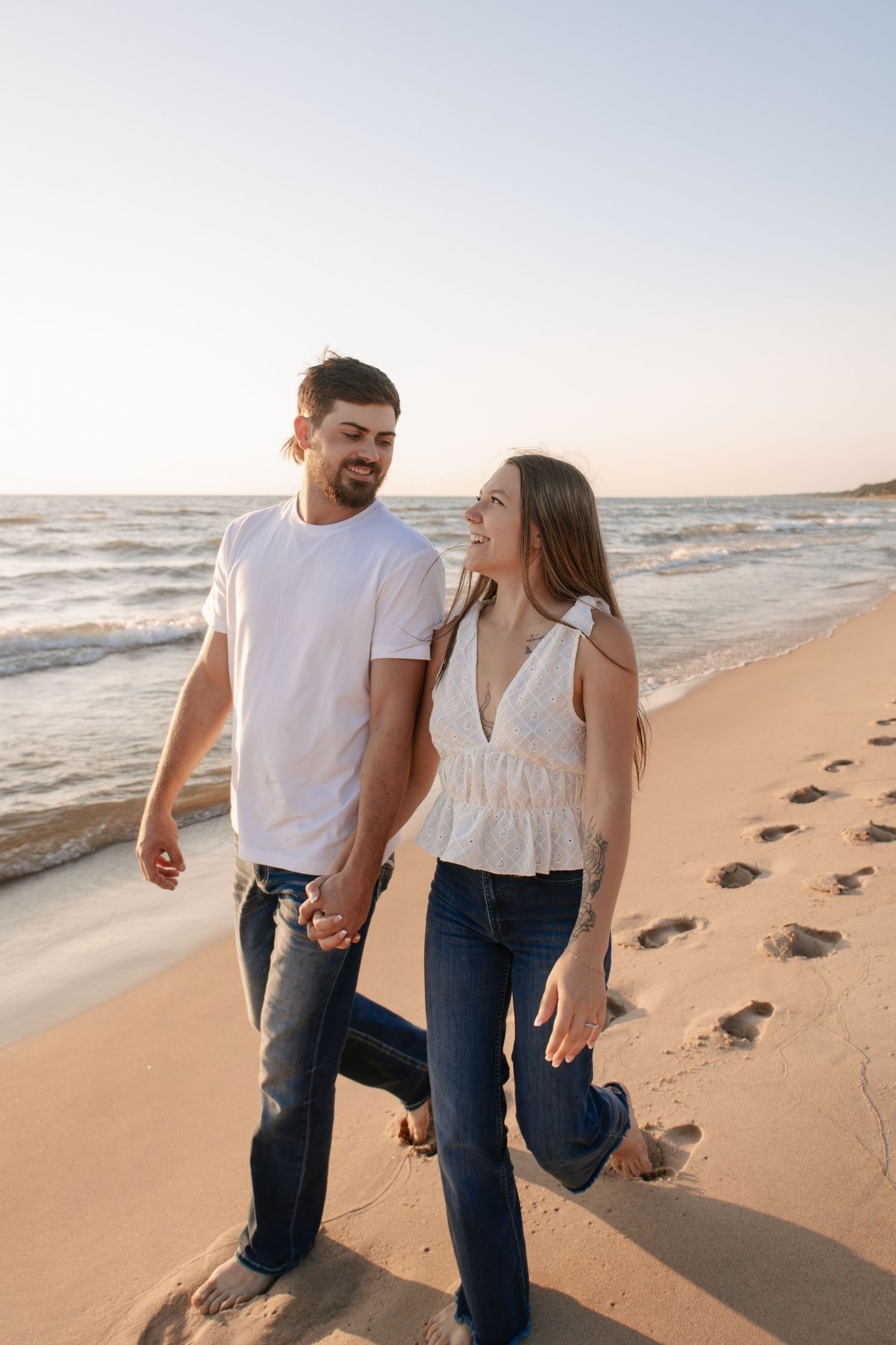 Happy couple holding hands walking barefoot on sandy beach at sunset, leaving footprints in the sand.