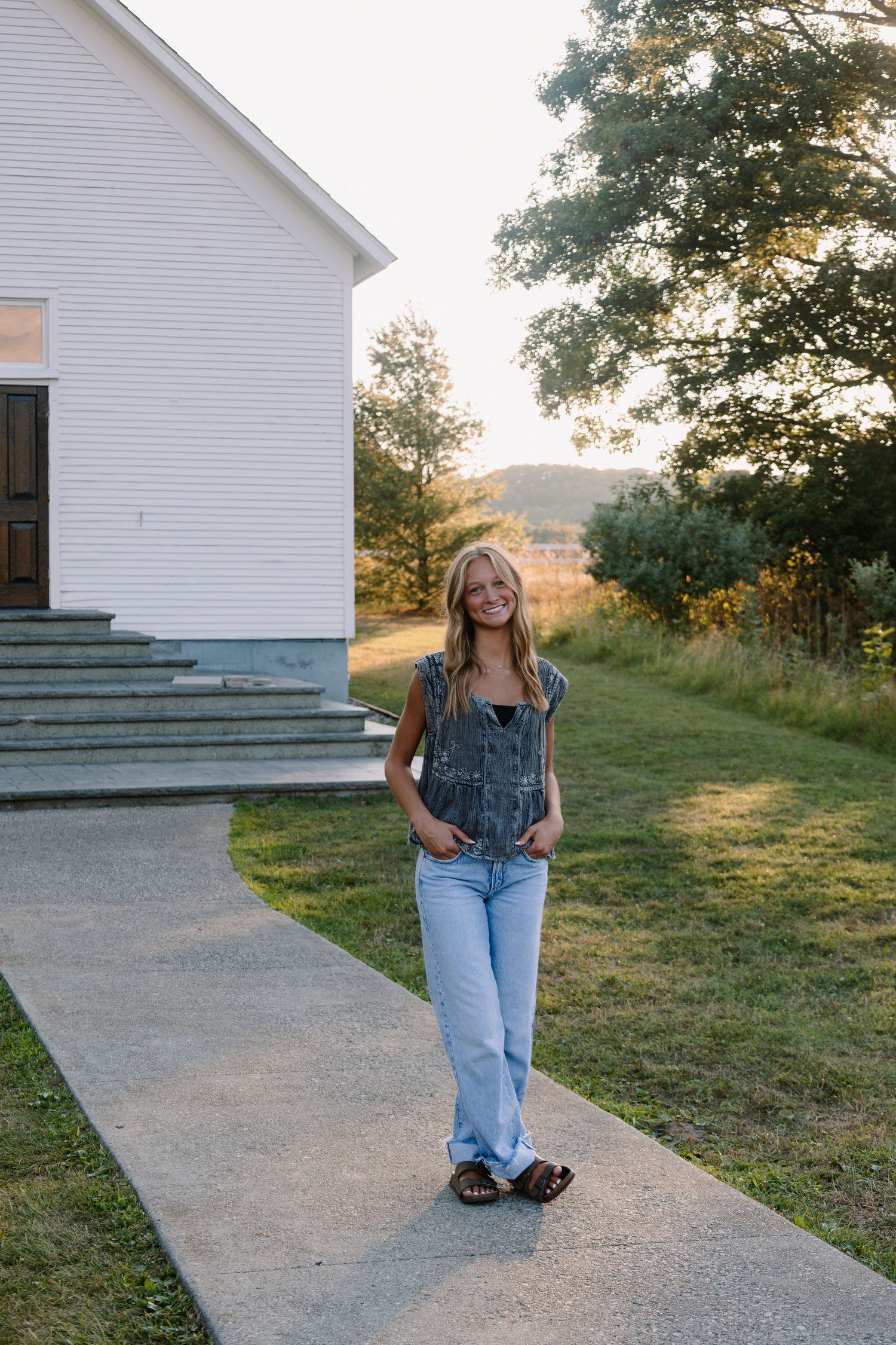Young woman in denim overalls and jeans smiles on a pathway beside a white building at golden hour.