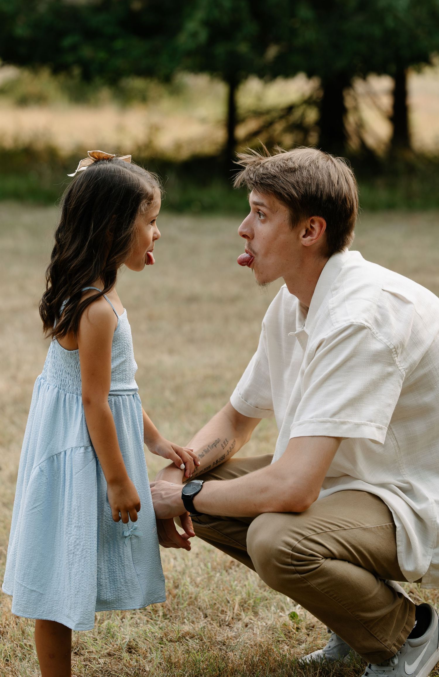 A young man crouches down sticking his tongue out playfully at a little girl in a blue dress outdoors.