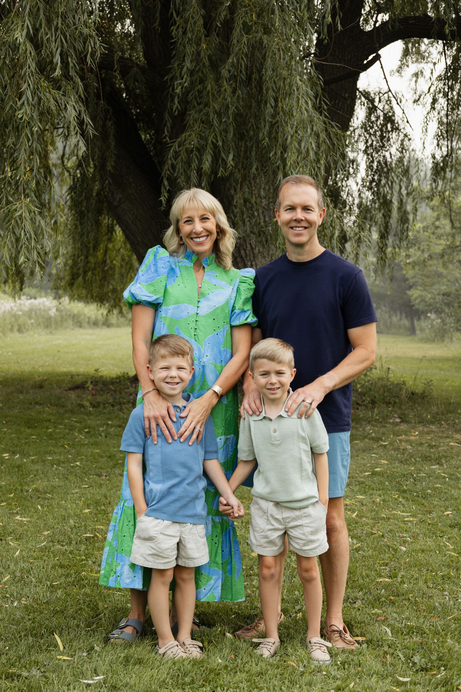 Family of four smiling outdoors under a weeping willow tree, parents with two young boys in coordinated outfits.