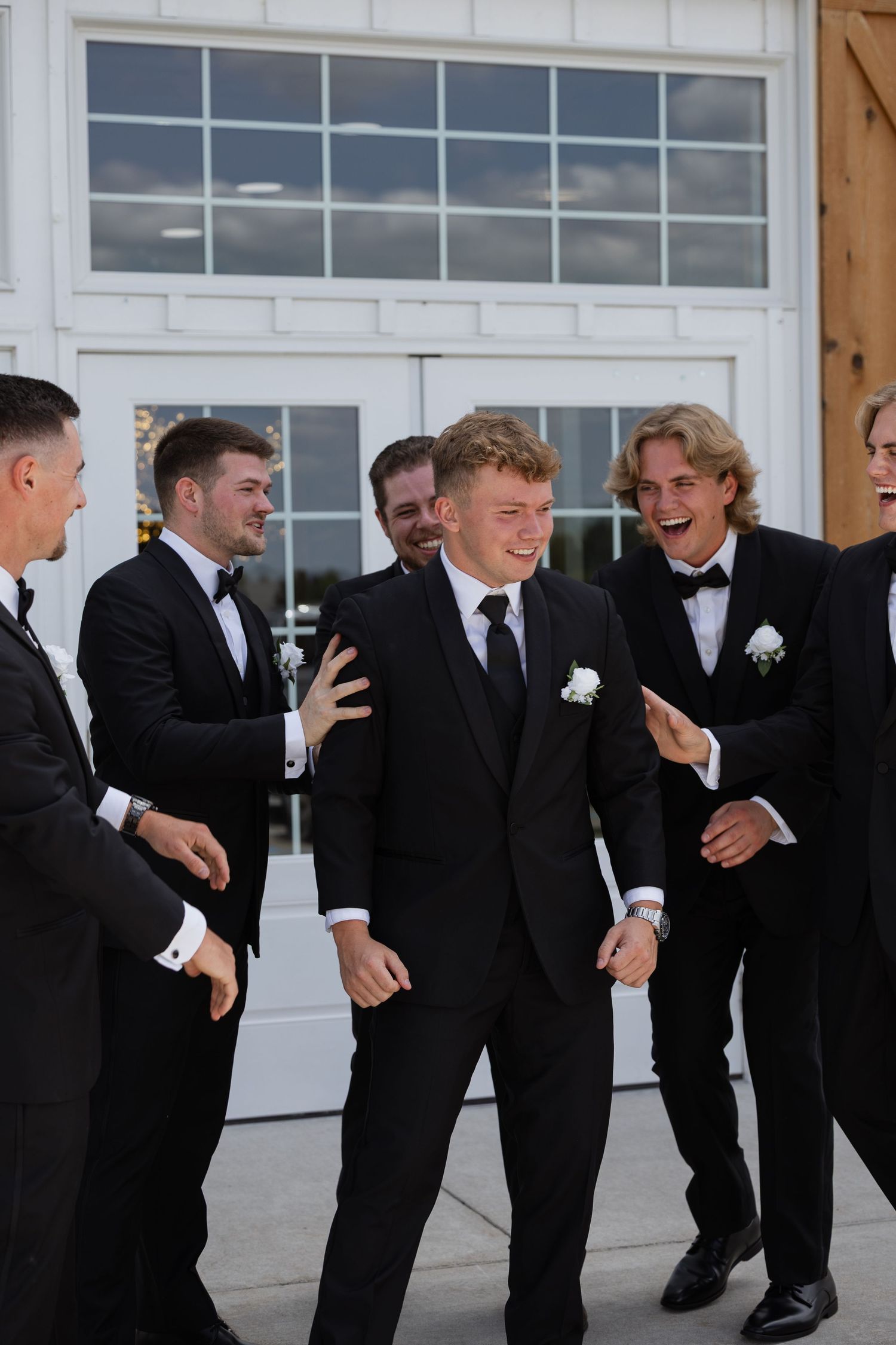 Groomsmen in black tuxedos laughing together outside white barn wedding venue on wedding day.