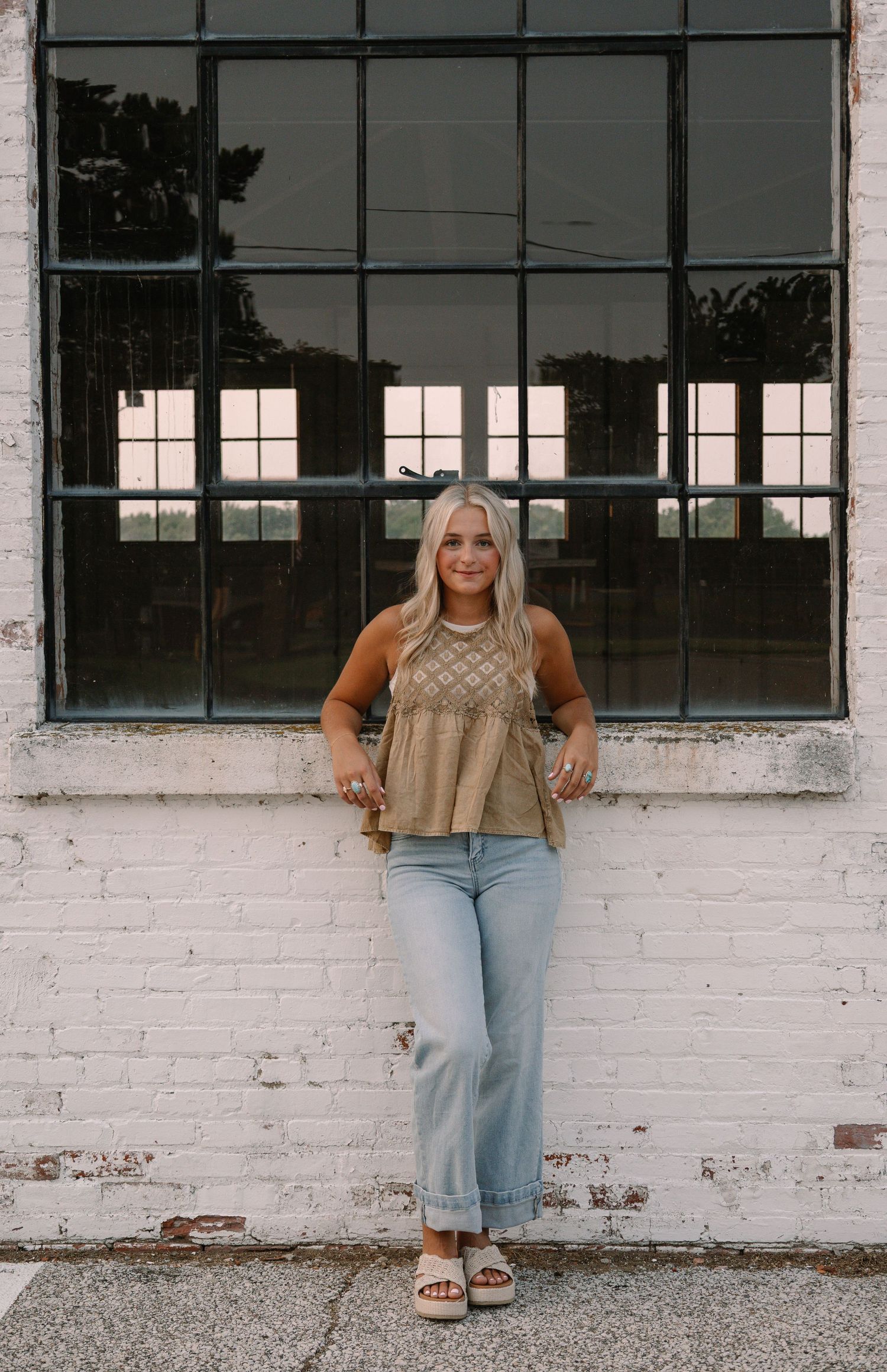 Woman in boho top and light jeans poses against white brick wall with large industrial window backdrop.