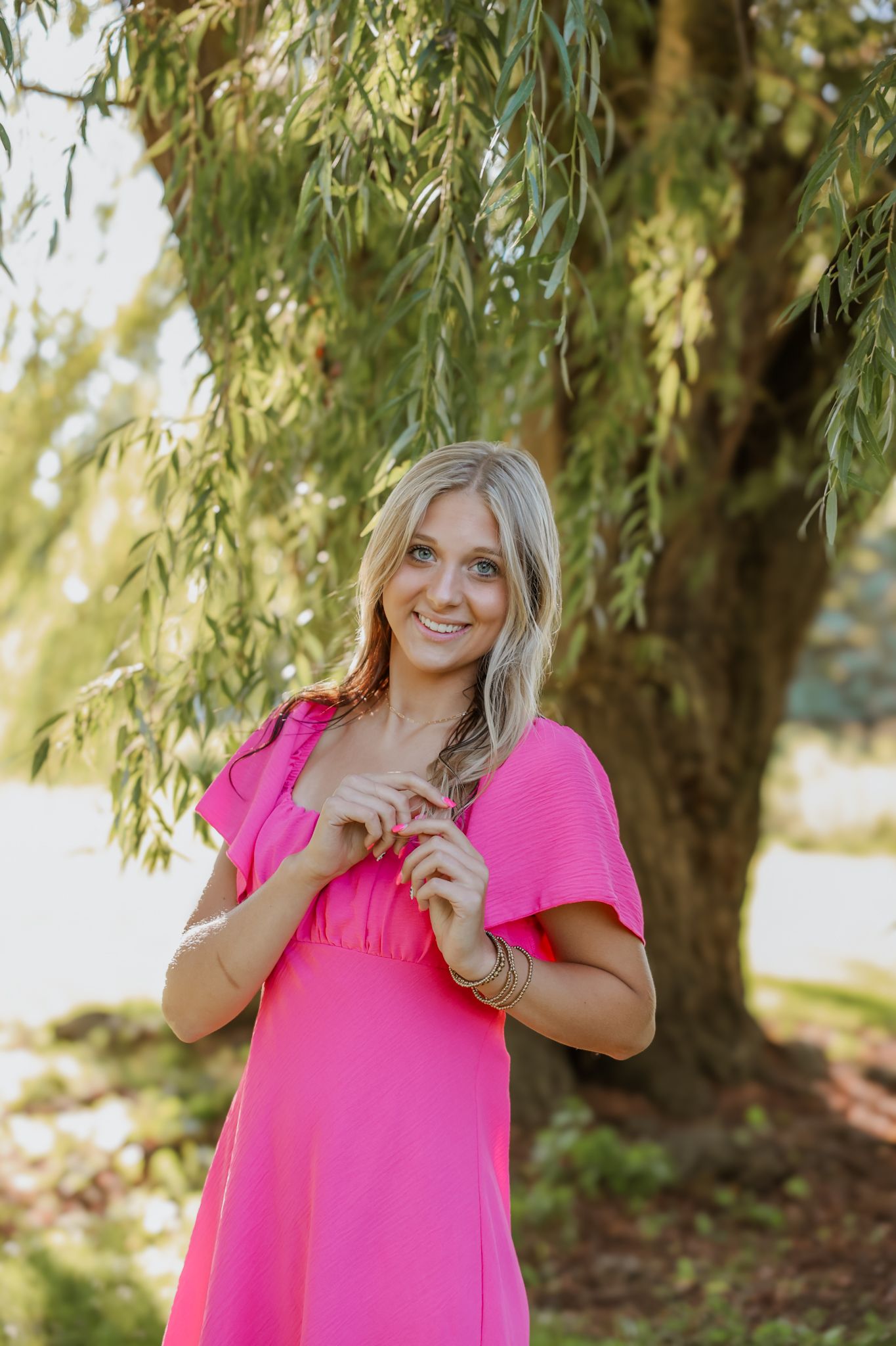 Smiling blonde woman in bright pink dress standing under a weeping willow tree outdoors.