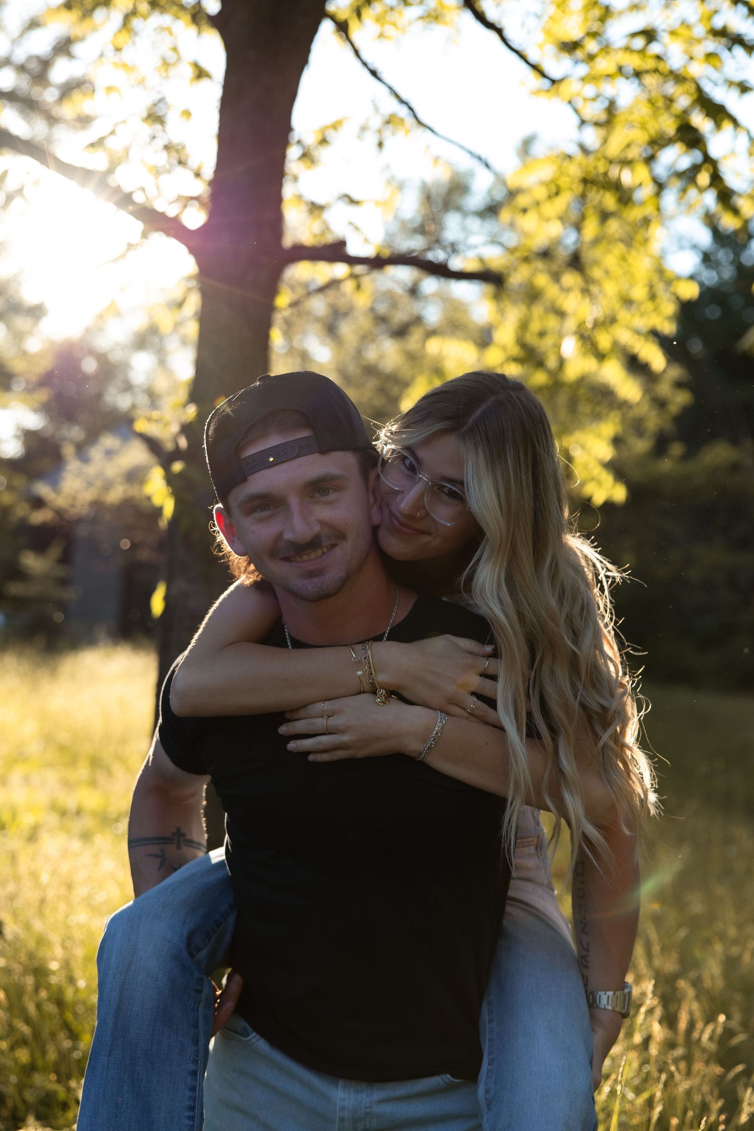 Happy couple sharing a piggyback ride in a sunlit forest, woman embracing man from behind with golden hour light.