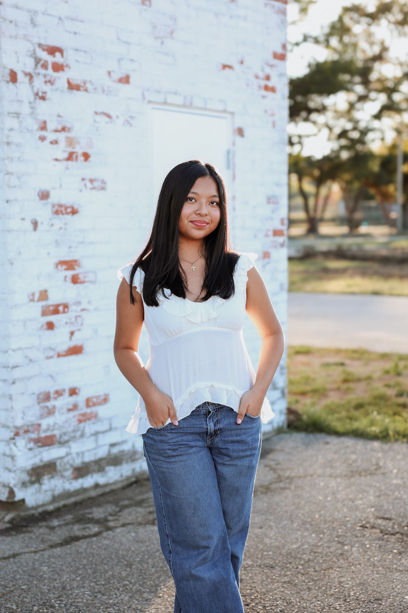 Young Asian woman in white ruffled top and jeans poses confidently by a white brick wall outdoors.