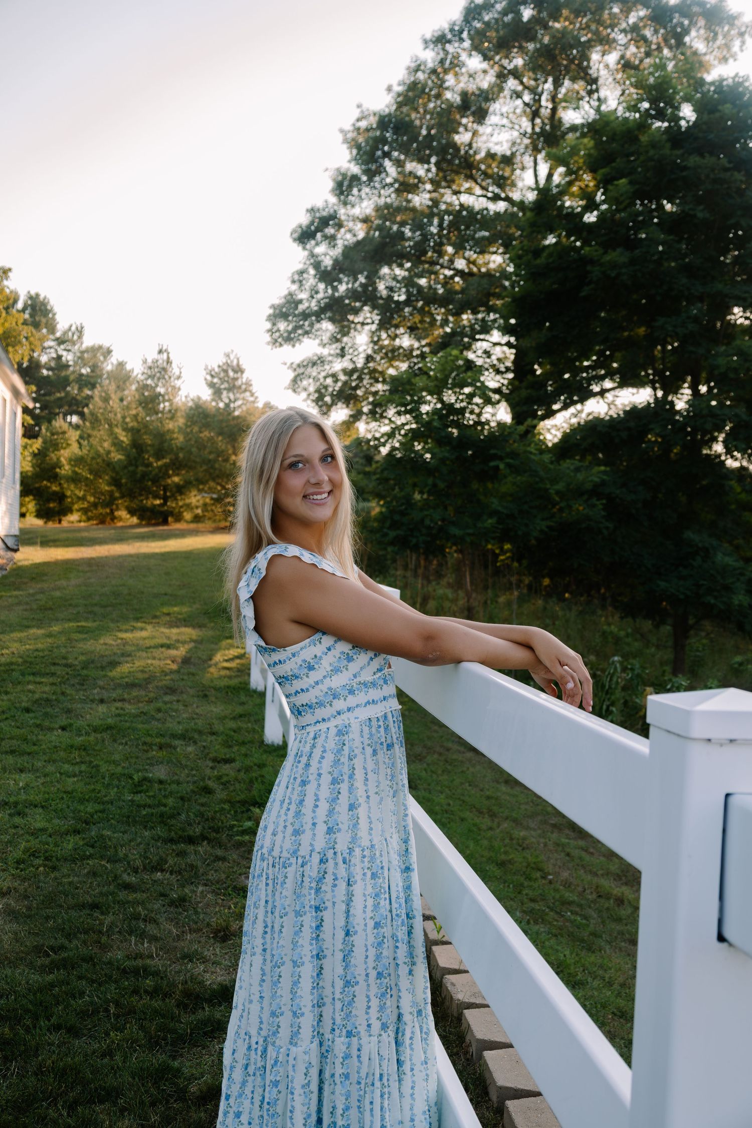 Smiling blonde woman in blue floral maxi dress leaning on white fence in lush green countryside at golden hour.