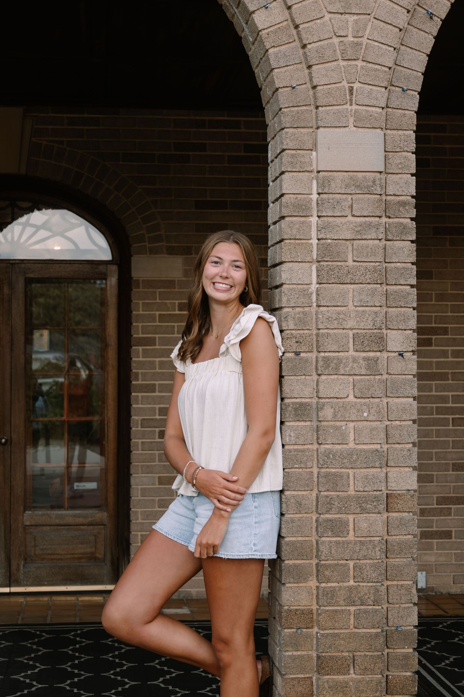 Smiling young woman in white top and denim shorts leaning against brick column near arched doorway.
