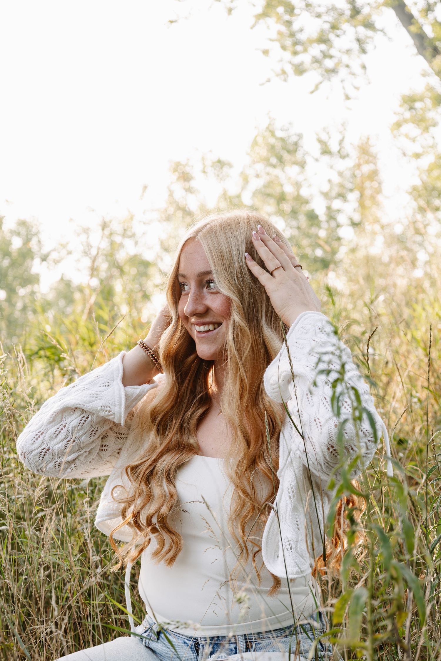Young woman with long wavy blonde hair smiling outdoors in a field, wearing a white lace cardigan and jeans.
