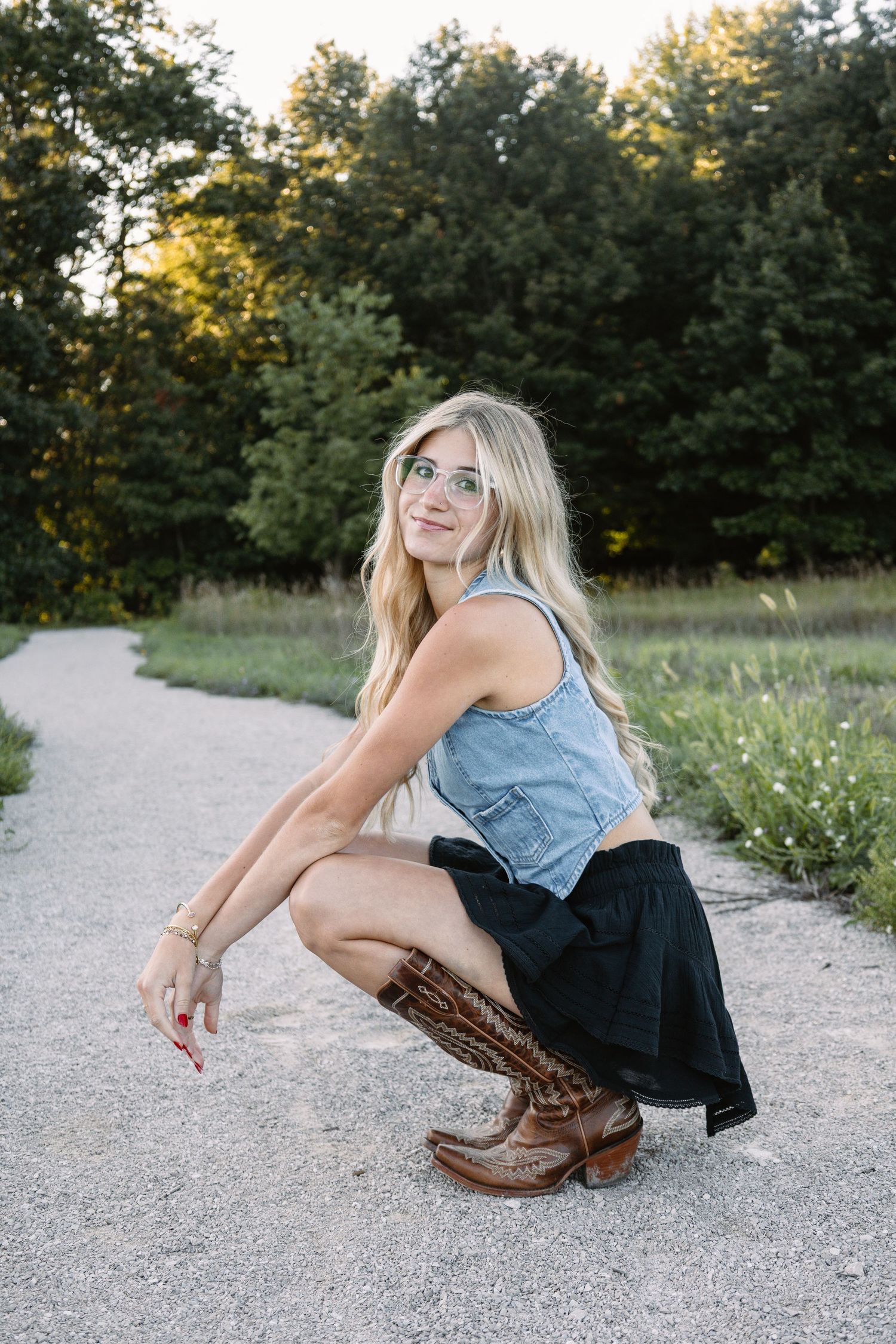 Blonde woman in denim top, black skirt, and cowboy boots crouching on a gravel path outdoors.