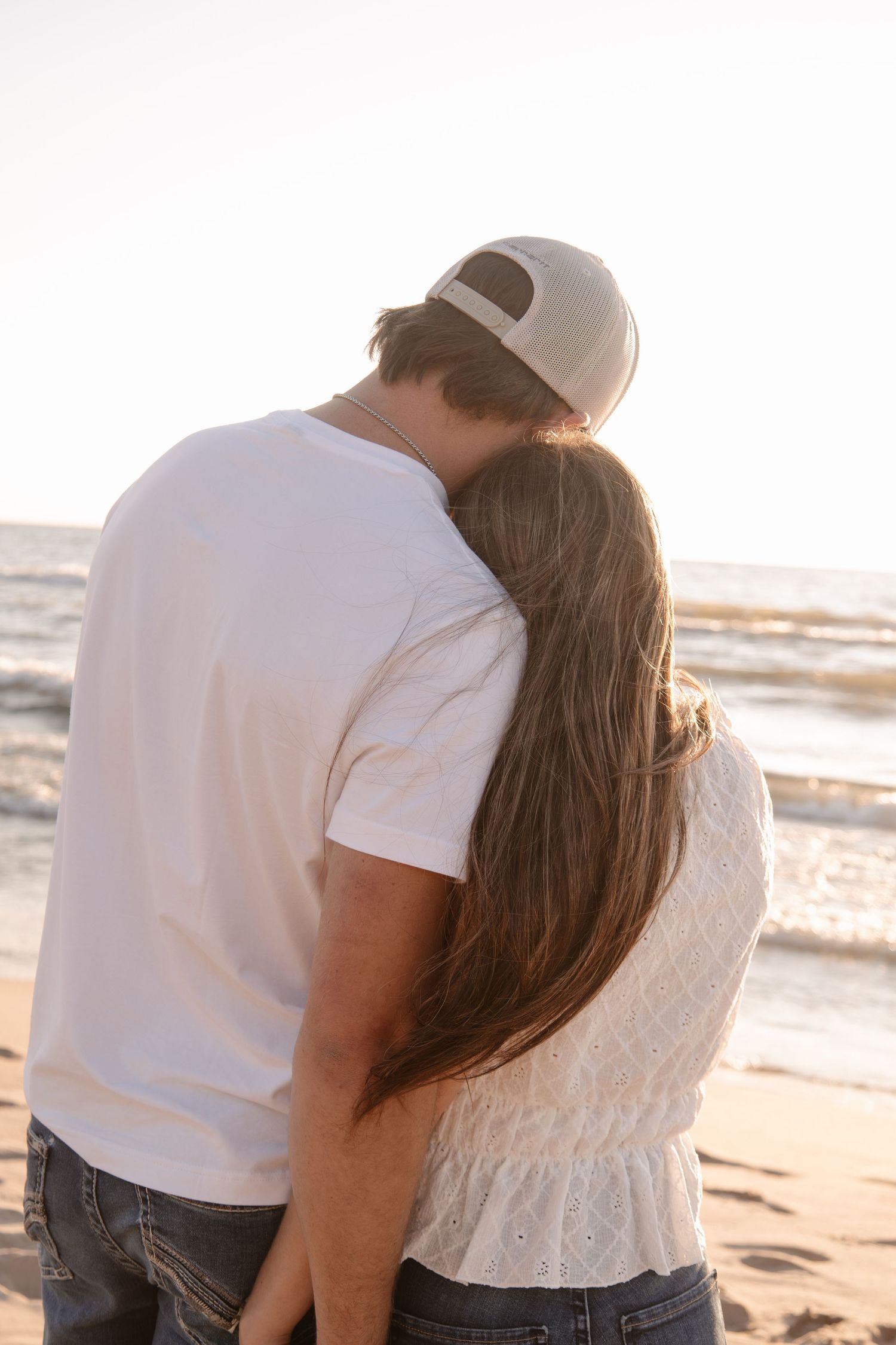 Couple embracing on a sandy beach at sunset, man in white tee and cap, woman with long brown hair.