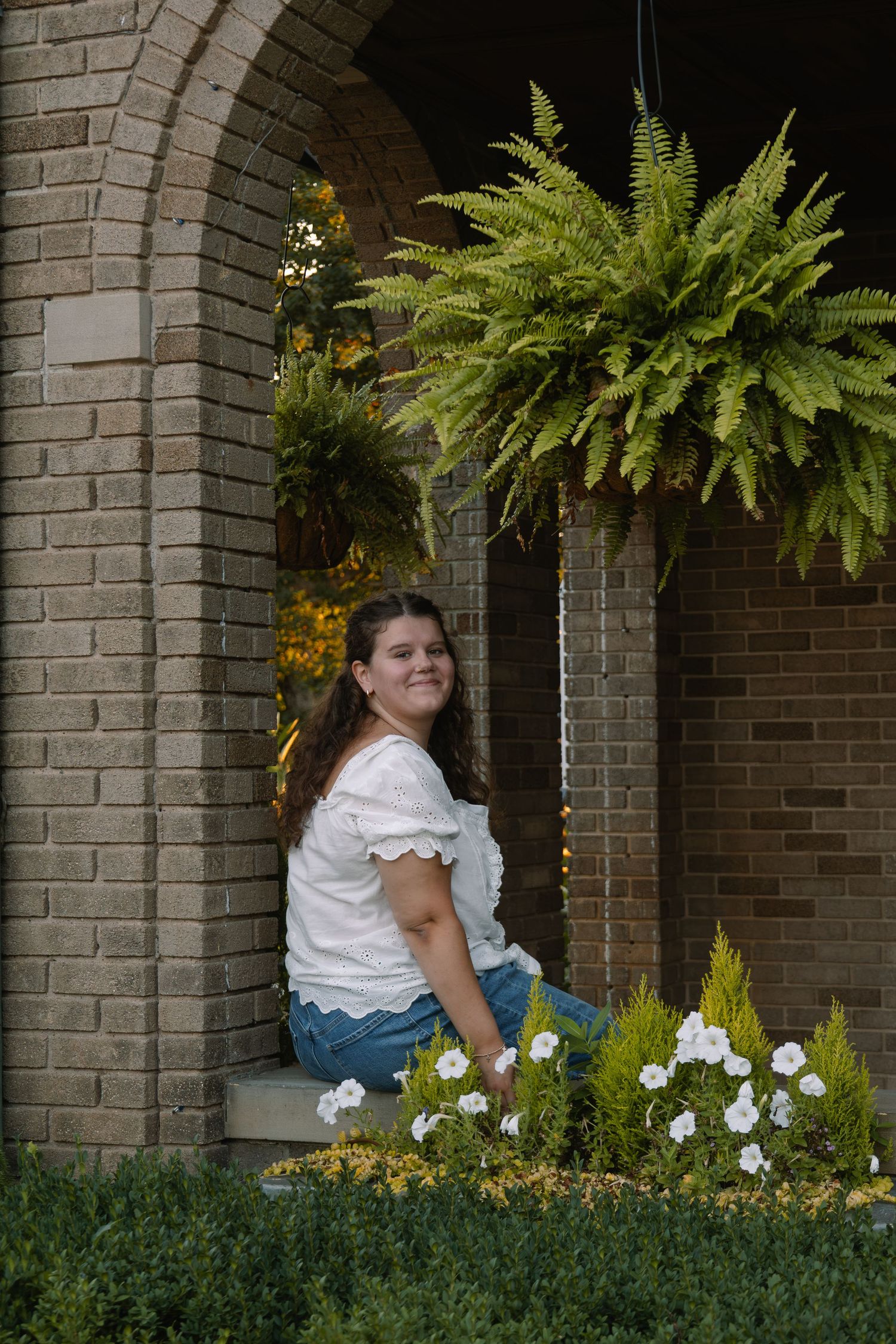 Young woman in white lace top sits smiling by brick archway with ferns and white flowers in garden setting.