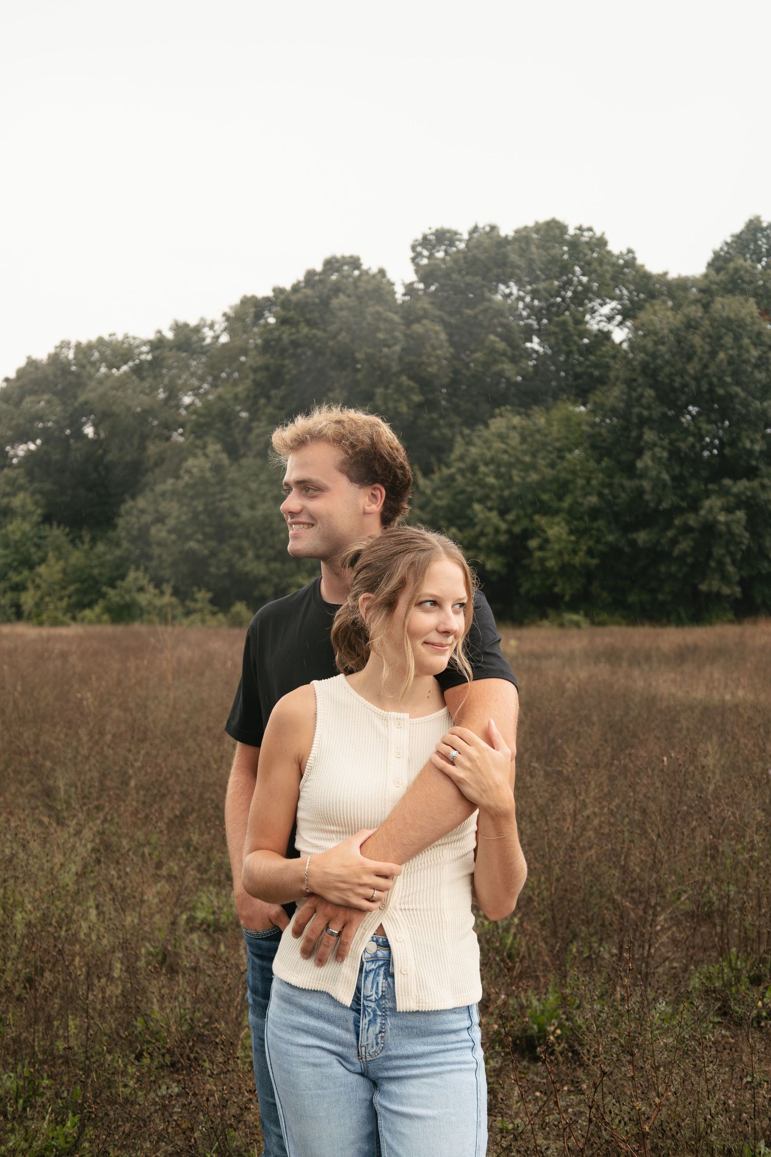 Young couple embracing in a field, man in black shirt hugging woman in white top and jeans, trees in background.