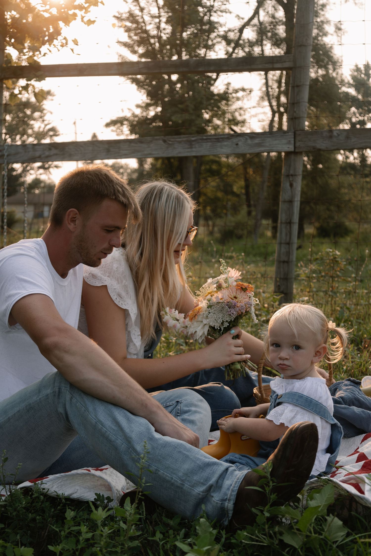 Young family sitting outdoors at golden hour, parents showing wildflower bouquet to toddler near rustic wooden fence.