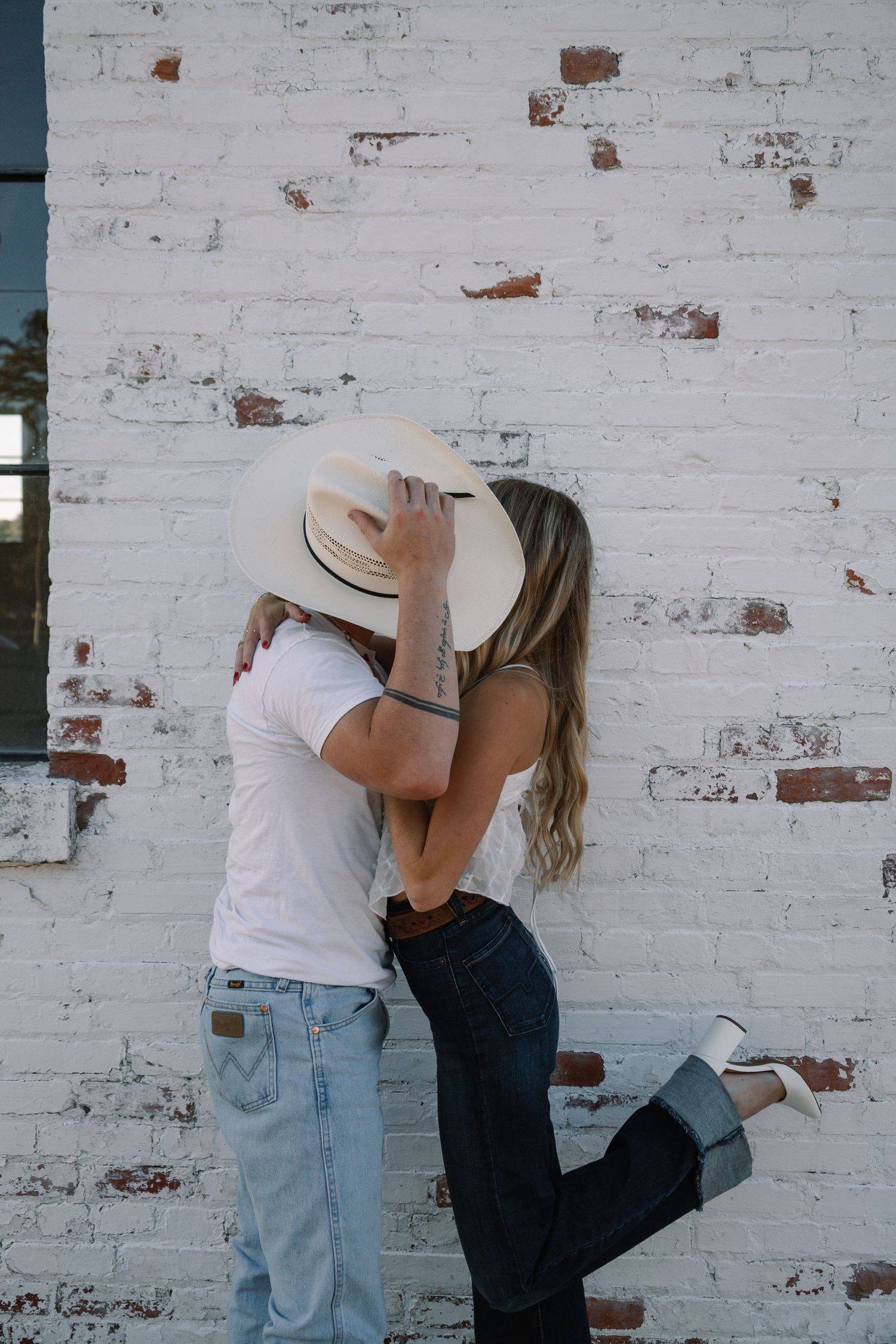 Couple sharing a romantic kiss hidden by a white cowboy hat against a rustic white brick wall outdoors.