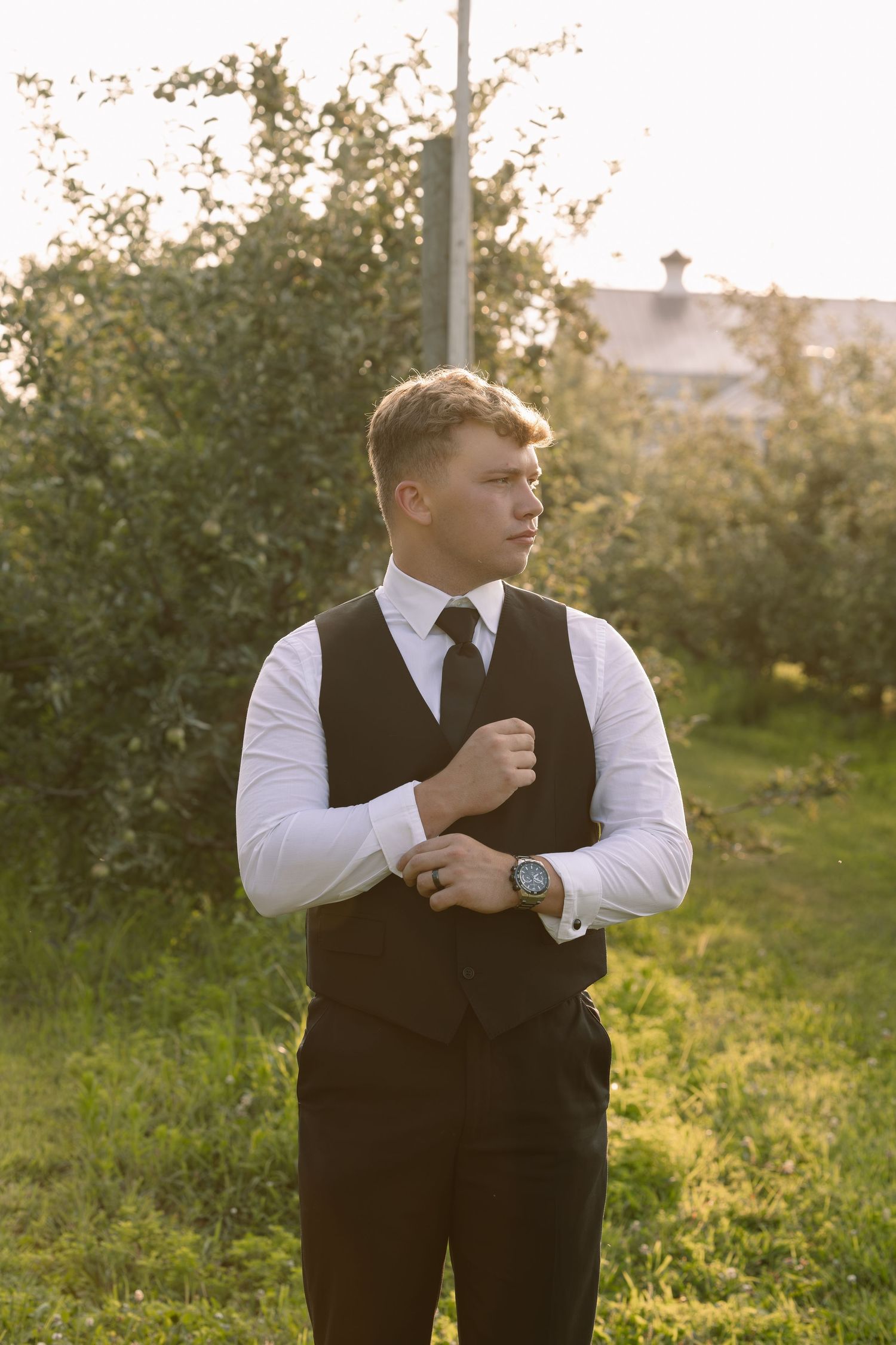Young groom in black vest, white shirt and tie adjusting cufflinks outdoors at golden hour.