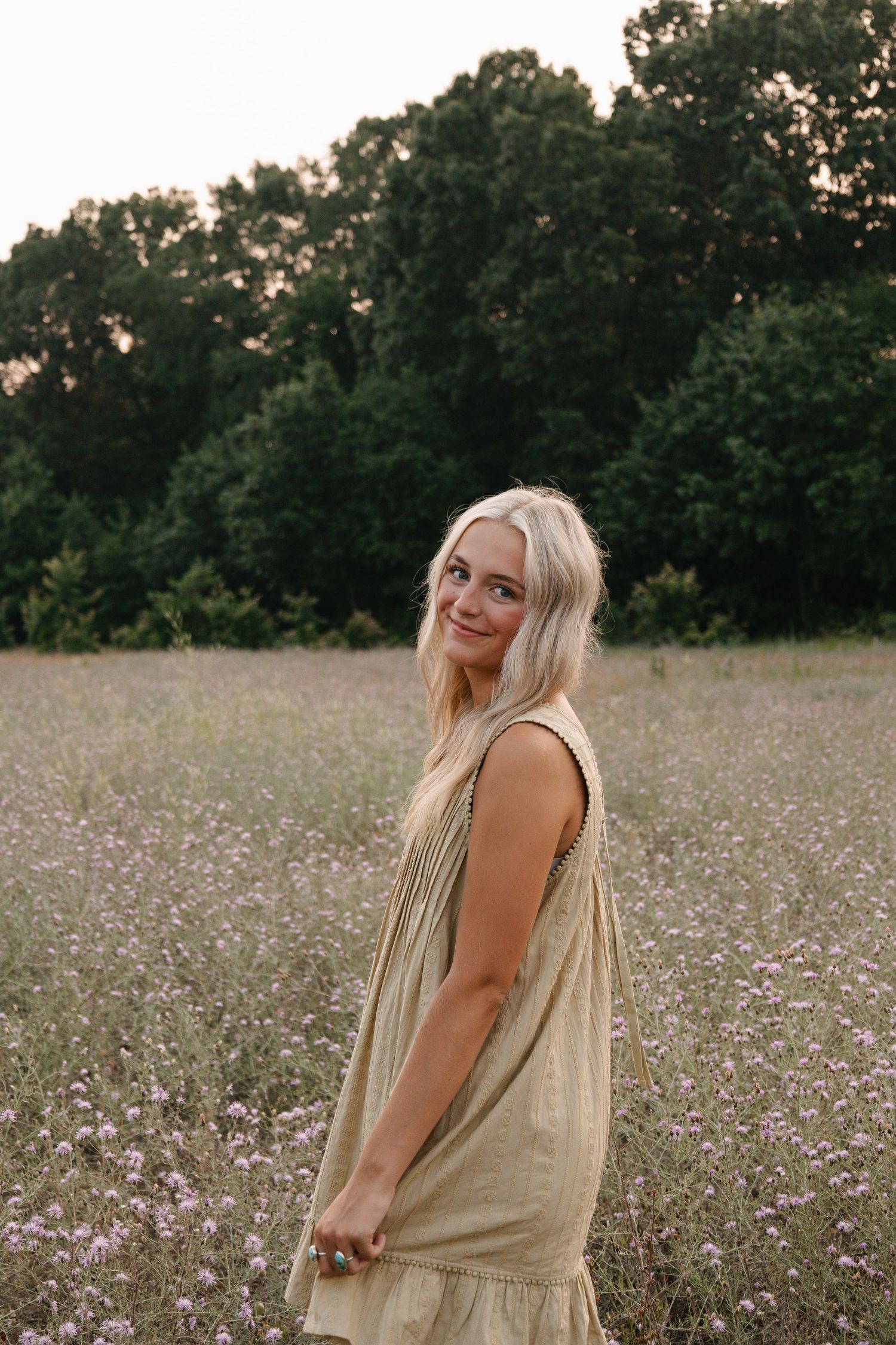 Blonde woman in a beige linen dress smiling in a wildflower meadow with green trees at golden hour.