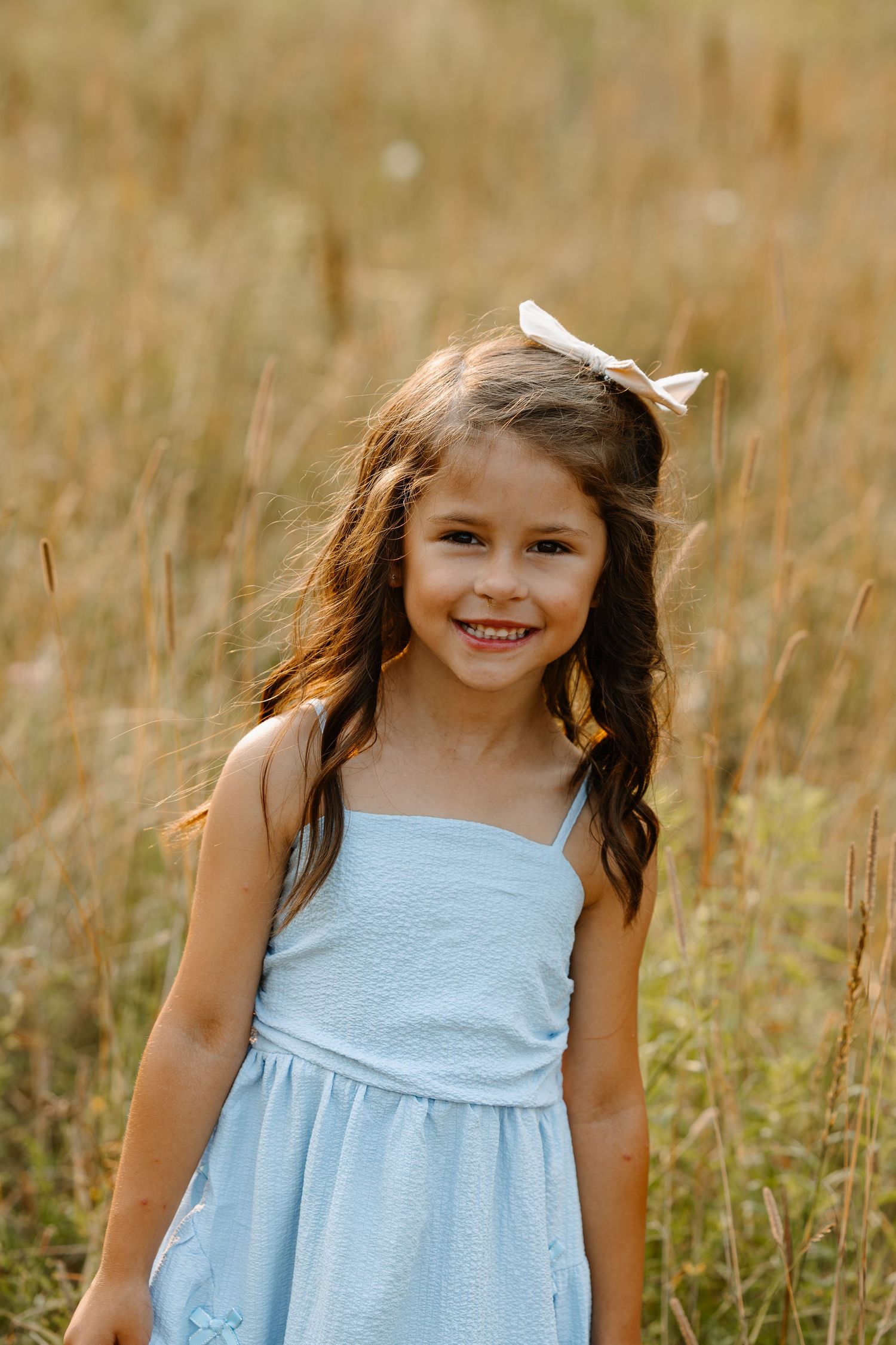 Smiling young girl with brown hair and white bow wearing a light blue sundress standing in a golden field.