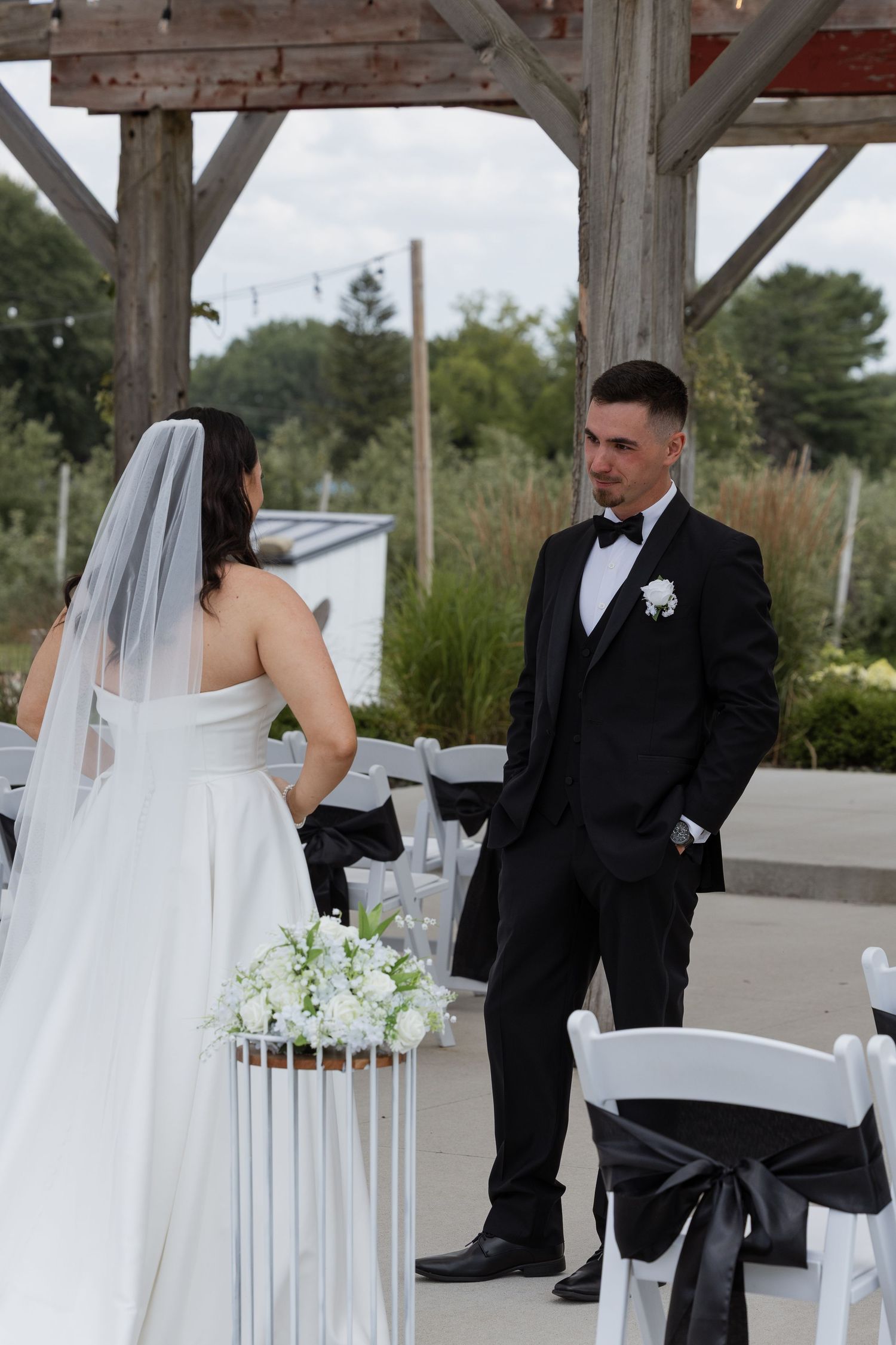 Groom in black tuxedo watches bride in white gown and veil at outdoor wedding ceremony.