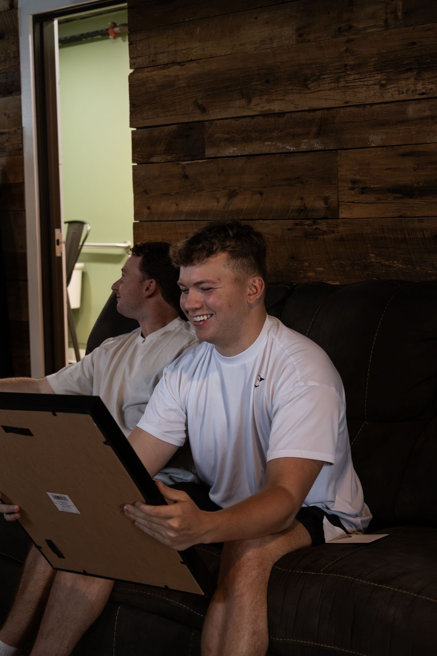 Young man in white t-shirt smiling while holding a framed picture, seated on a dark couch indoors.