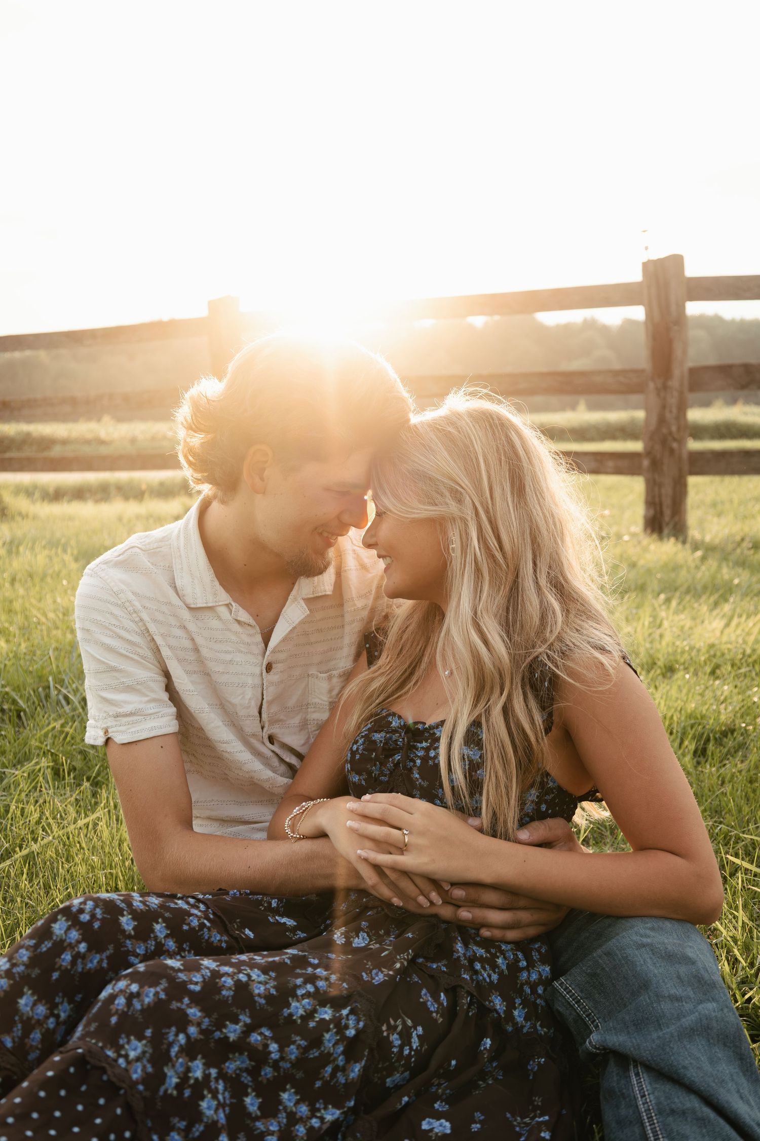 A couple sits in a sunlit field, foreheads touching romantically, with a wooden fence and golden sunset behind them.