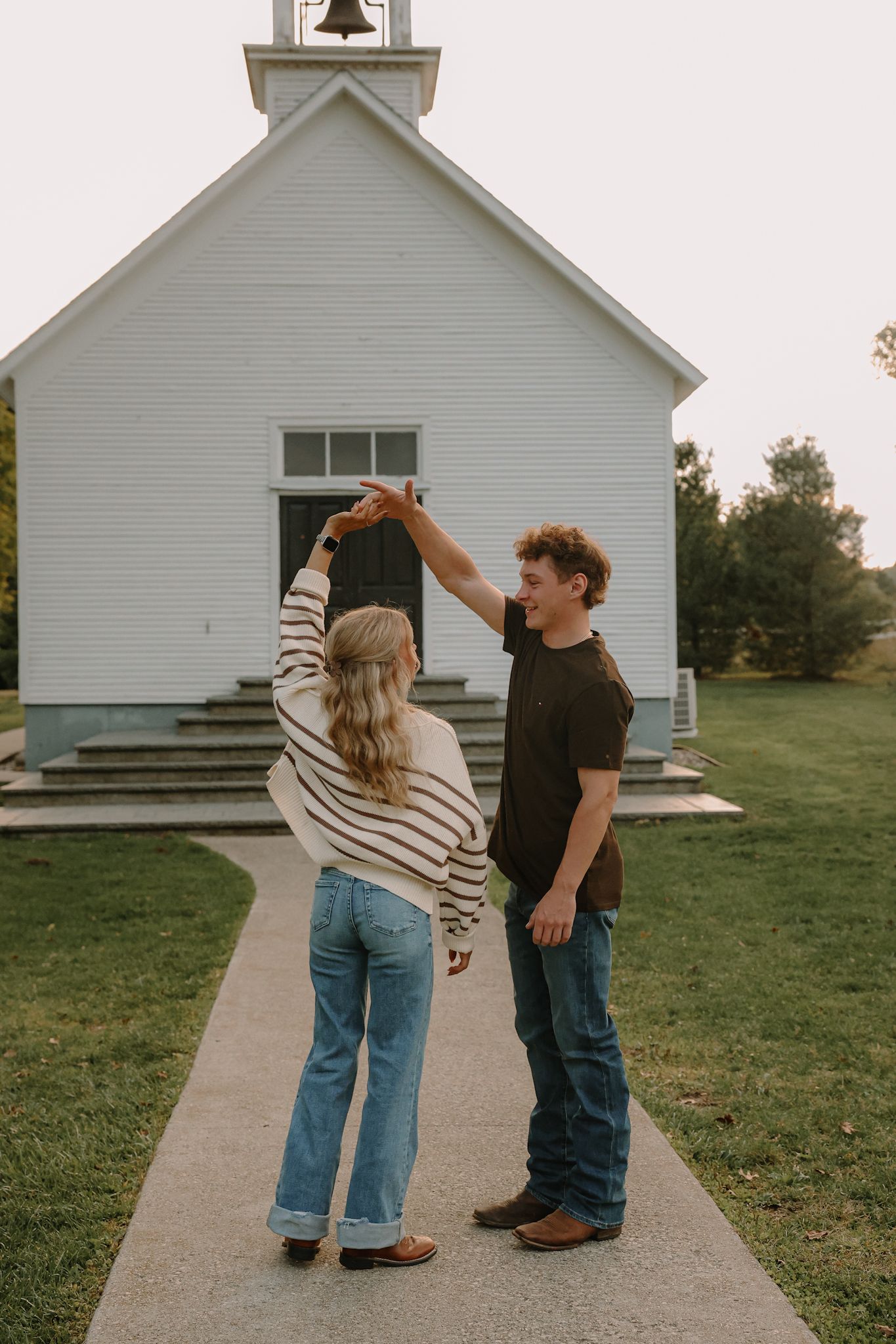 A couple dances playfully on a pathway in front of a charming white country church at dusk.