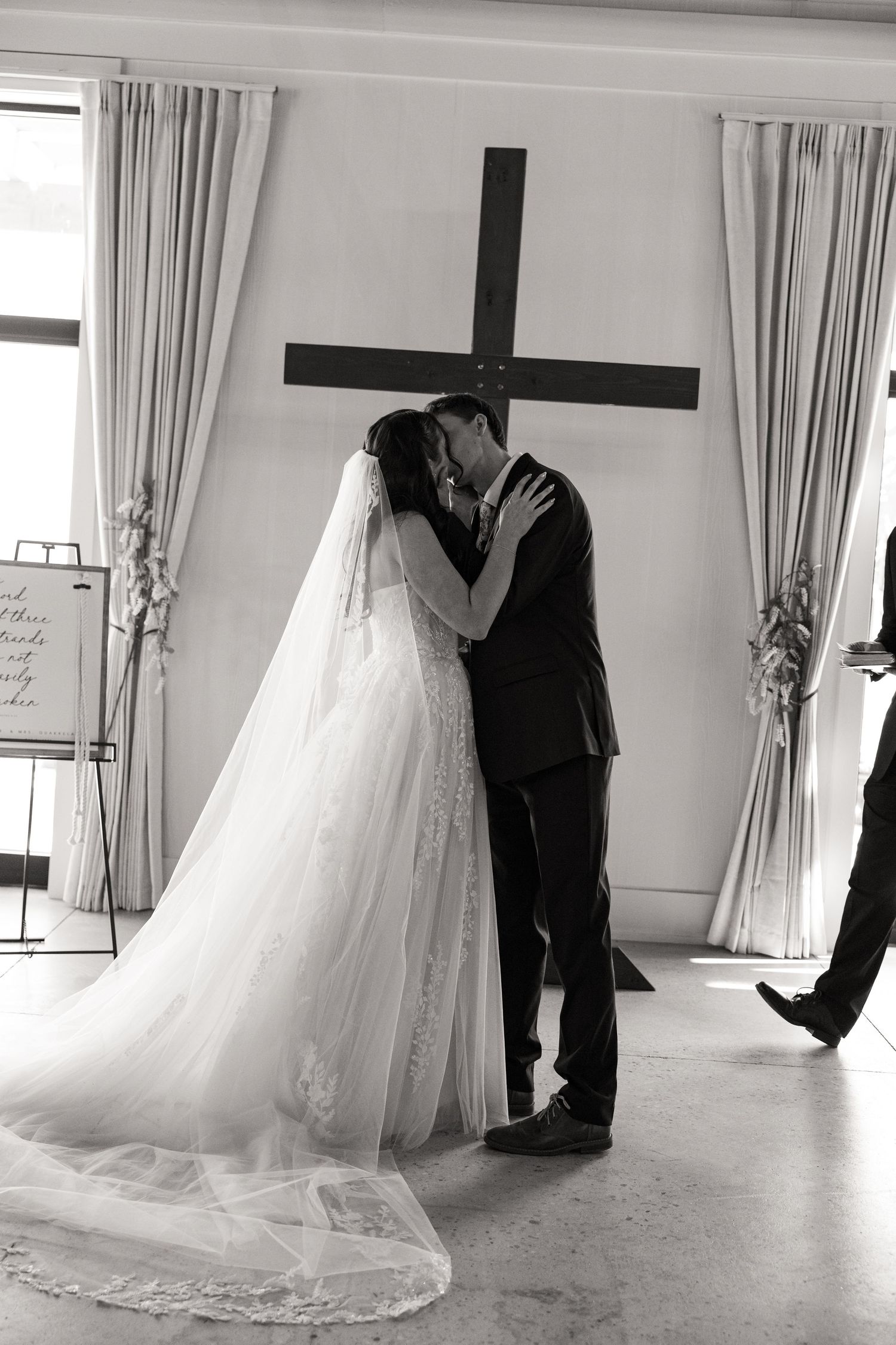 Black and white photo of bride and groom sharing first kiss in front of wooden cross.