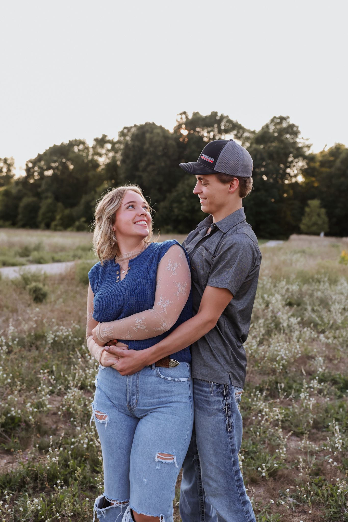A couple embraces in a wildflower field at sunset, the woman in a blue top smiling up at the man in a gray shirt.