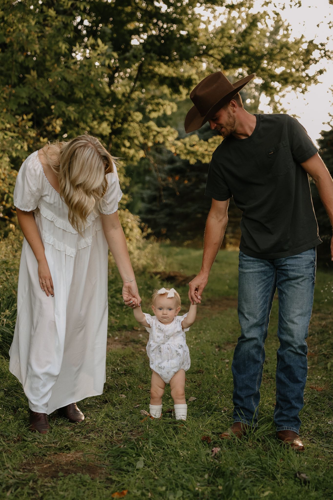 Parents in golden hour light helping their baby girl walk on grass, dad wearing brown cowboy hat.